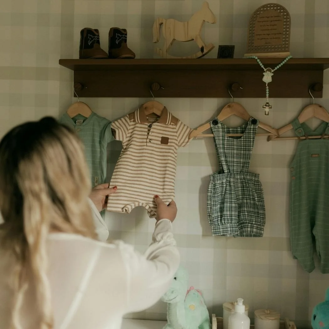 A woman shopping for baby clothes in a store, holding a beige and white striped baby onesie in front of other baby clothes on the wall, with a pair of brown shoes and decorative items on a shelf above.