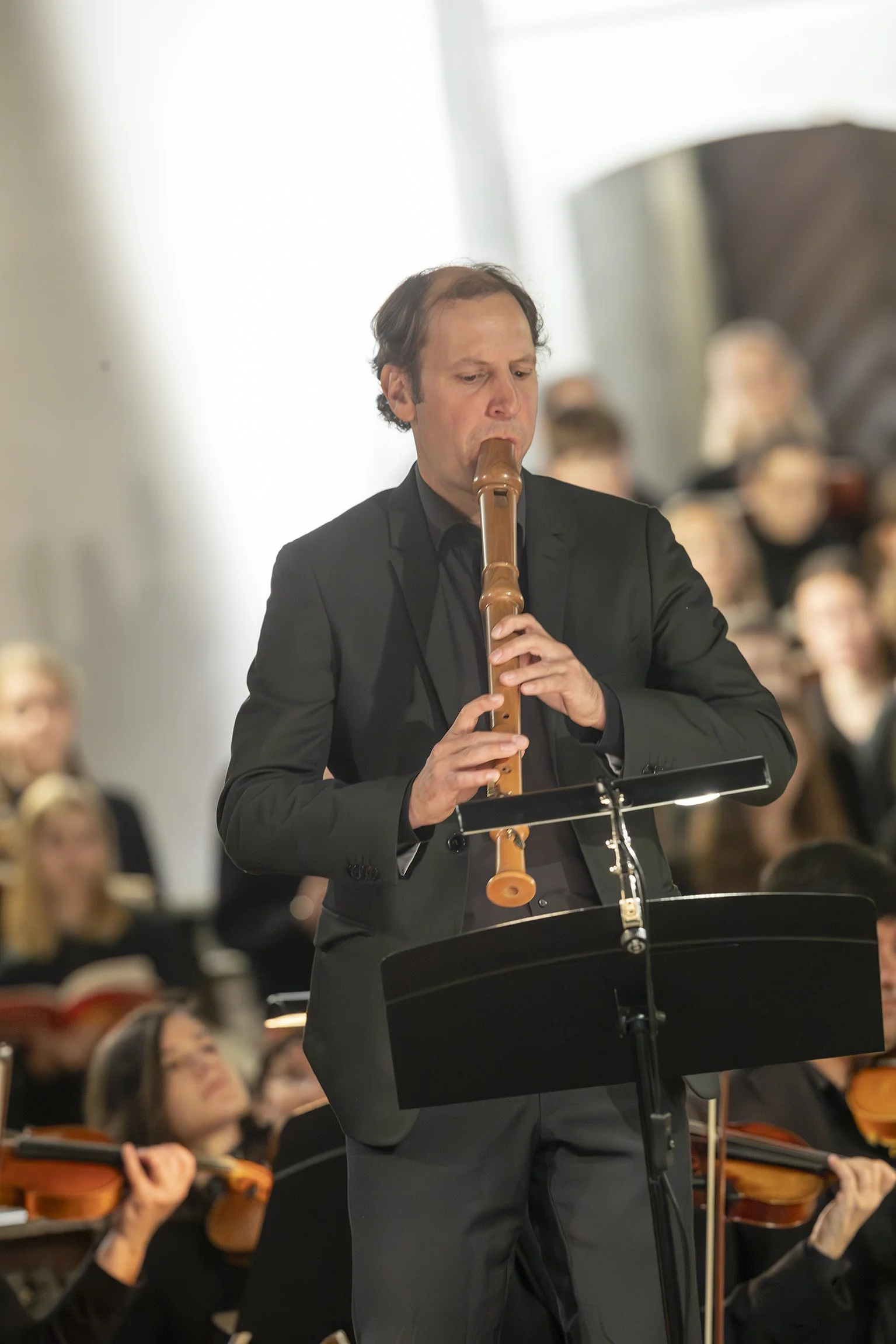 Benedikt Dreher spielt in einem Orchester solistisch eine Blockflöte. Im Hintergrund sind Musiker mit Violinen zu sehen.