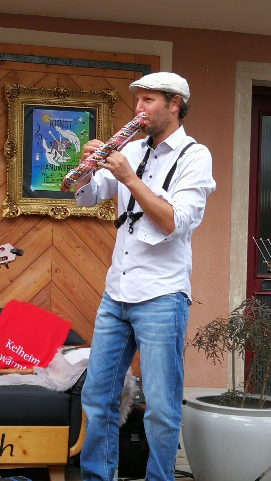 Ein Mann spielt auf einer Melodica, trägt ein weißes Hemd, eine graue Mütze und Jeans, im Hintergrund ein Kunstposter und eine Holz-Wand.