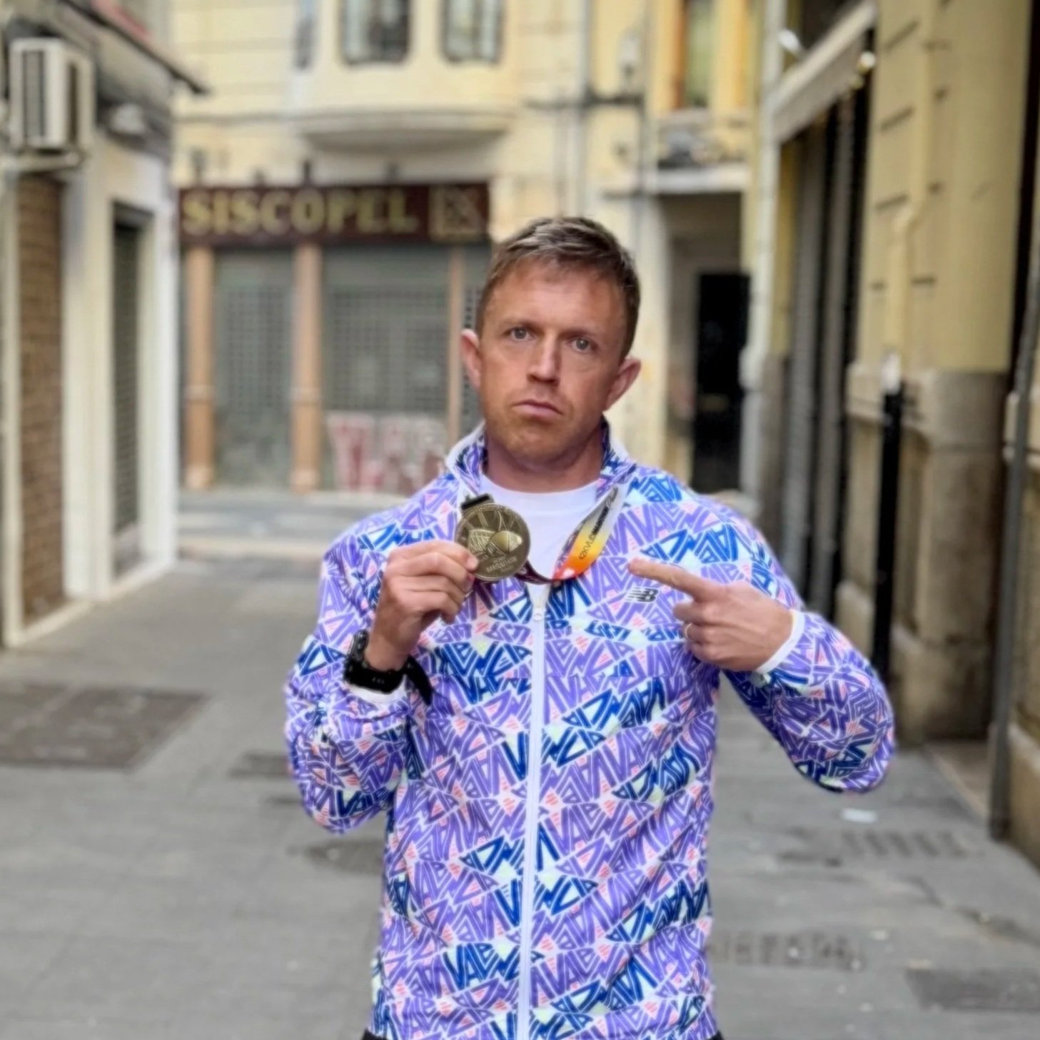 Man in colorful Valencia Marathon jacket holding a Valencia Marathon medal and pointing at it on a city street