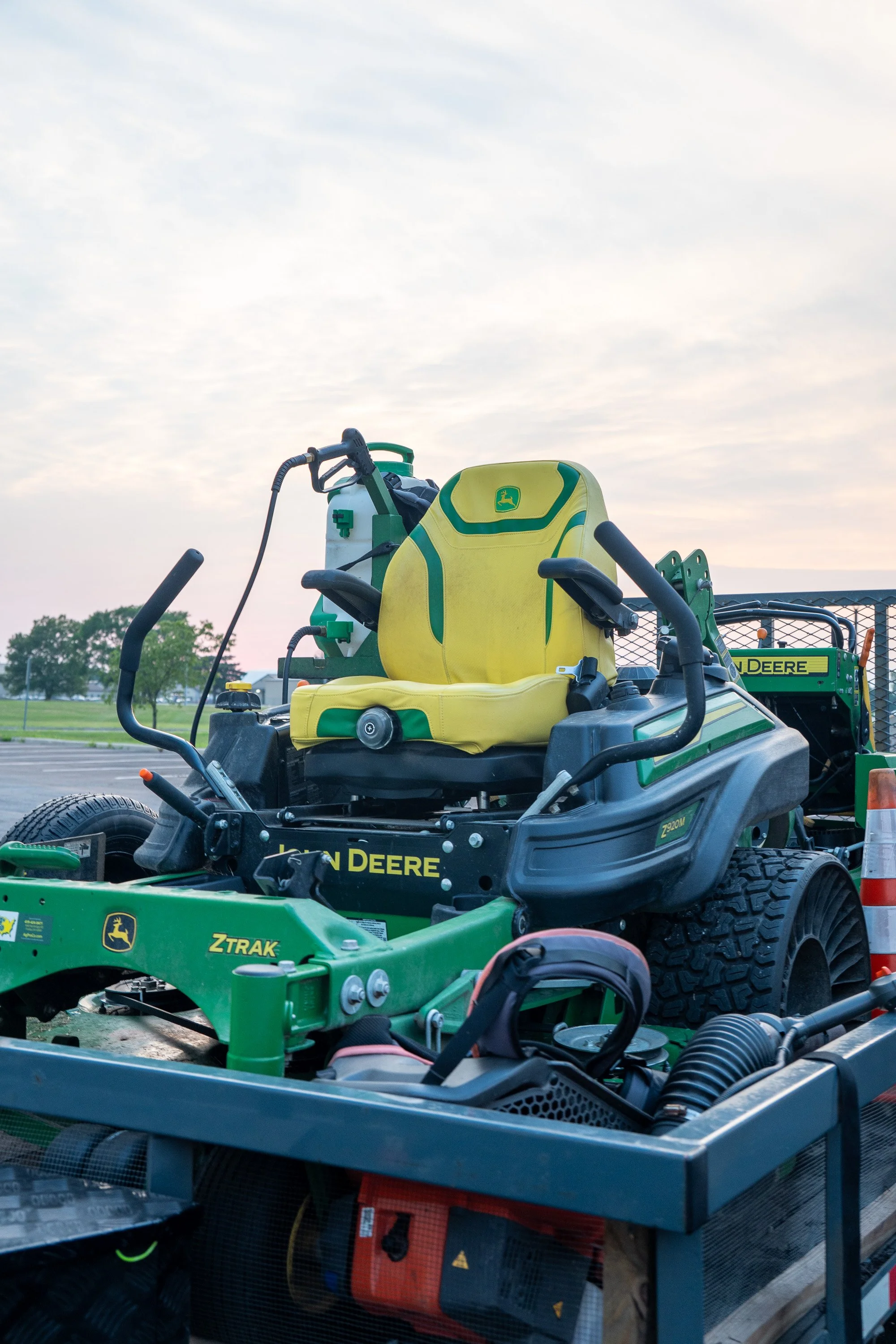 Various John Deere lawn care equipment, including a yellow and green ride-on mower and a green ZTRAK mower, on a trailer at a park during daytime with a background of trees and a cloudy sky.