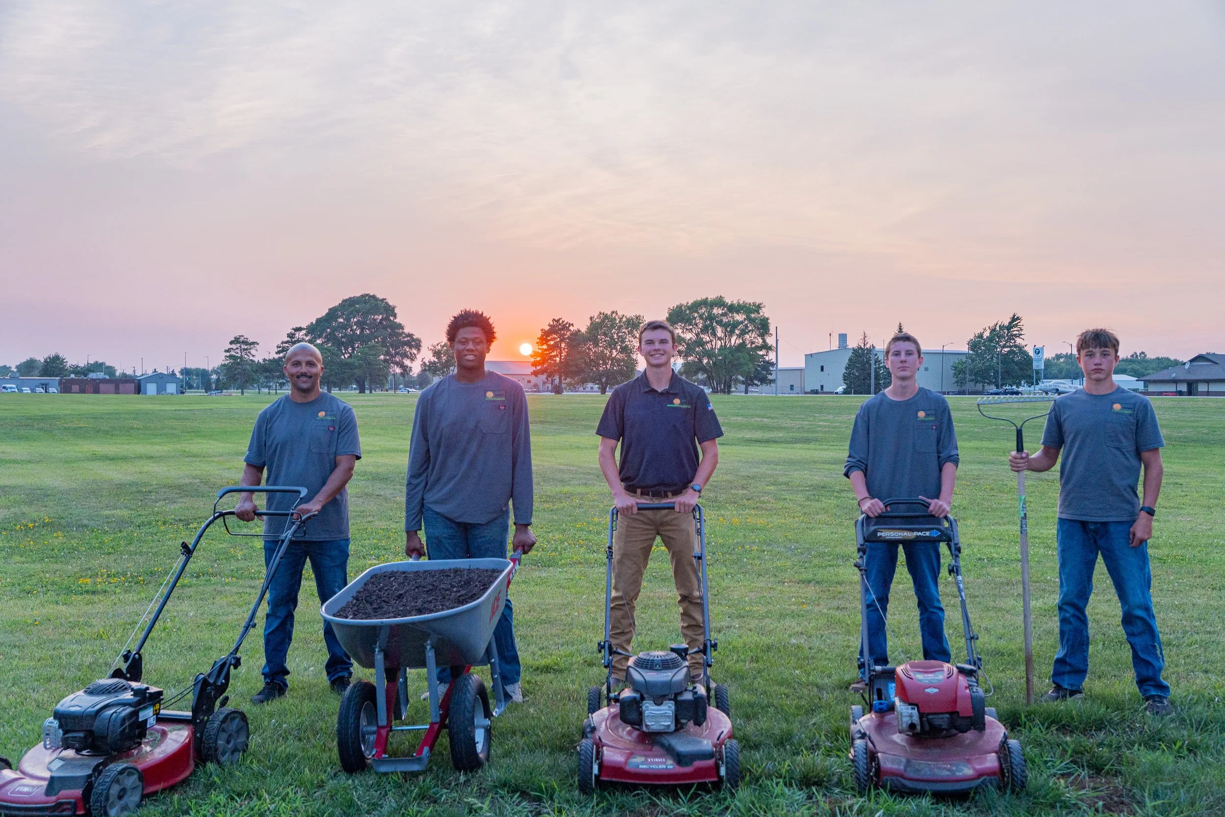 A group of five people standing on a grassy field at sunset, each holding or standing next to lawn equipment including lawnmowers and a weed trimmer.