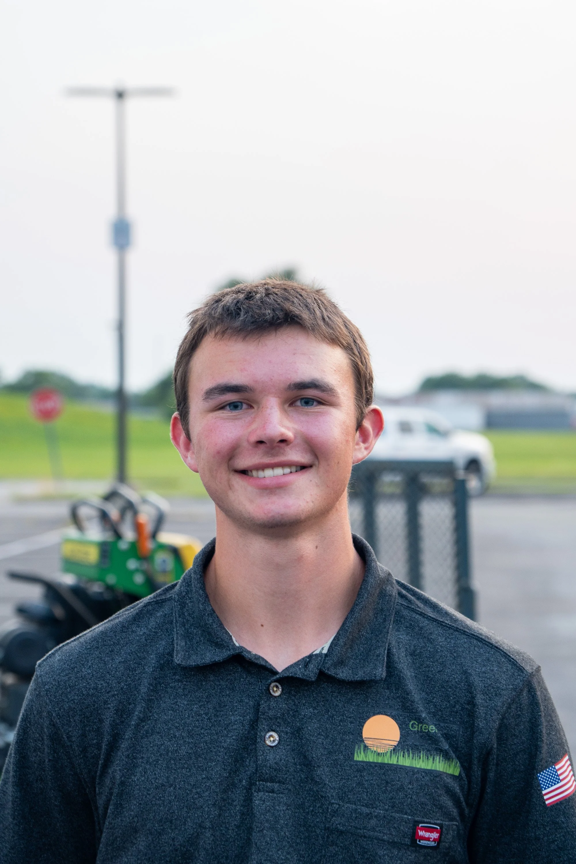 A young man with short brown hair and blue eyes smiling outdoors during daytime, wearing a dark gray collared shirt with patches including an American flag and a logo for Green.