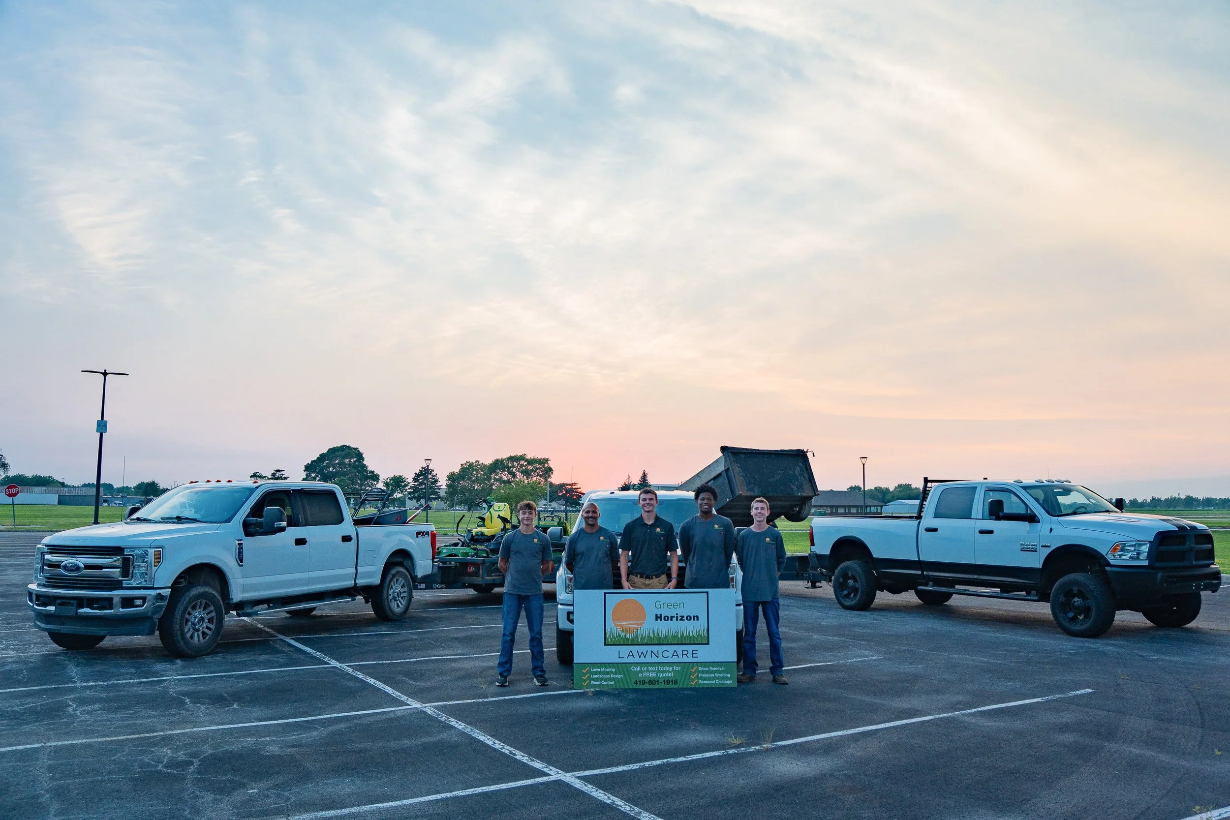 A group of five men standing in a parking lot, posing behind a sign that reads "Green Horizon Landscaping." They are flanked by a white pickup truck on the left and a black pickup truck on the right, with lawn care equipment and a trailer in between. The sky above is partly cloudy at sunset.
