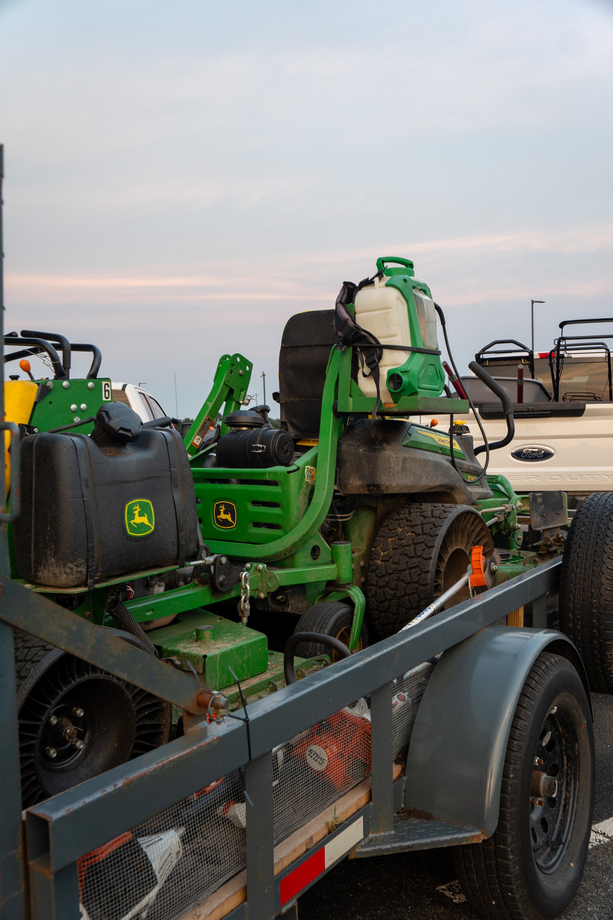 Green John Deere lawn mower on a trailer, with a white pickup truck and other equipment in the background, under an overcast sky.