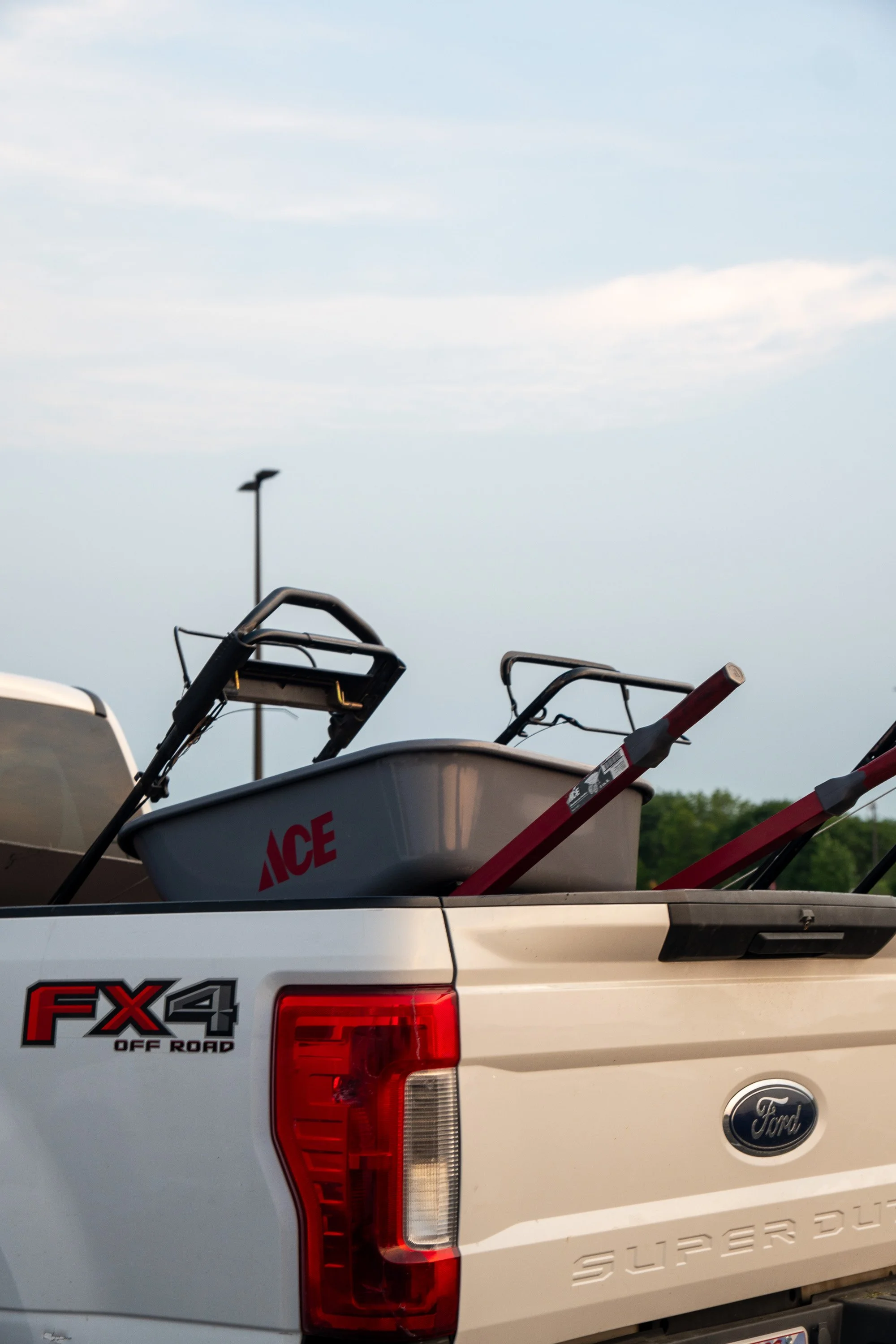 The image shows the back of a white Ford Super Duty pickup truck loaded with boat accessories, including boat seats and a ladder, with the Ace Hardware logo visible on the boat and a streetlamp in the background.