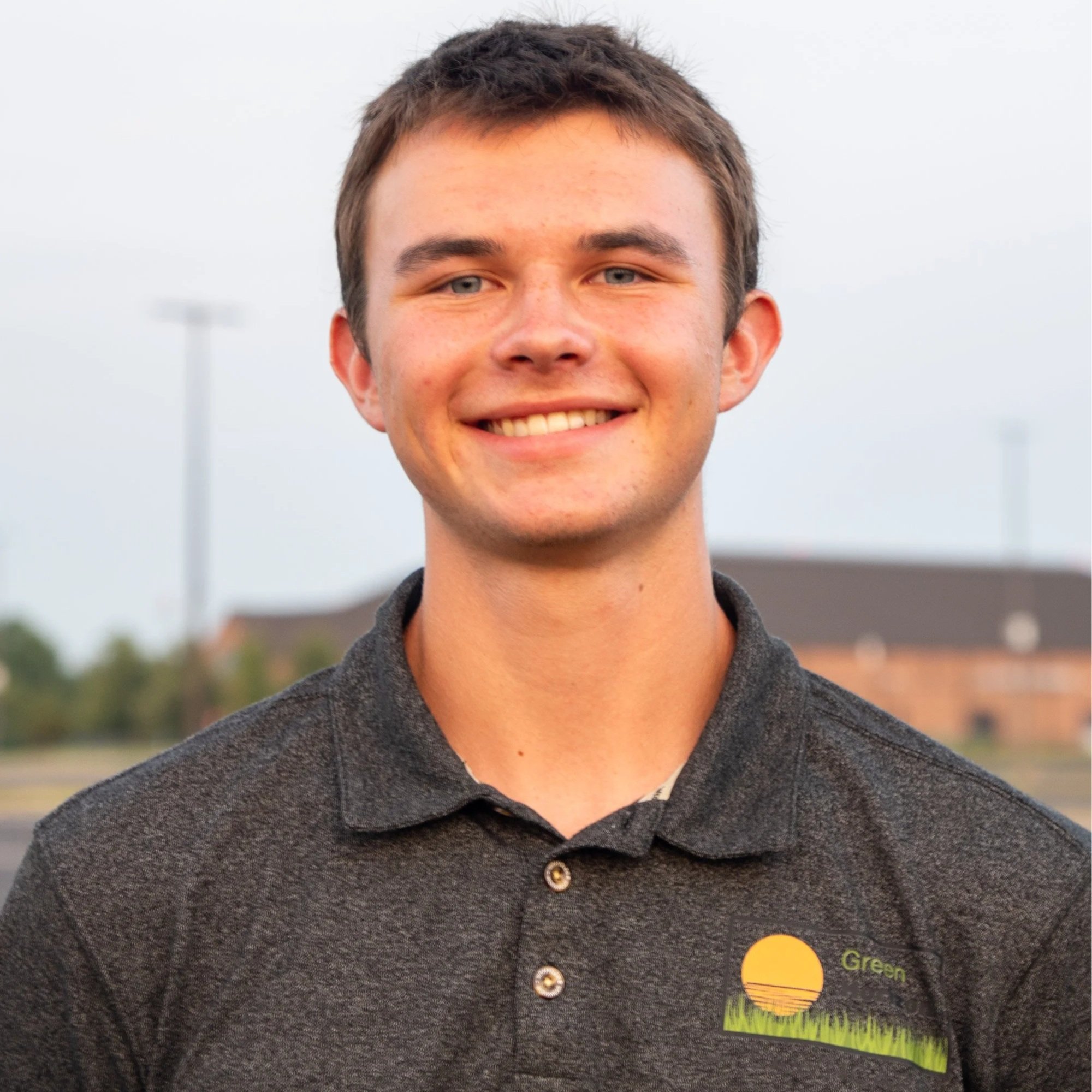 Close-up of a young person's face with short brown hair and blue eyes, smiling outdoors on a cloudy day.