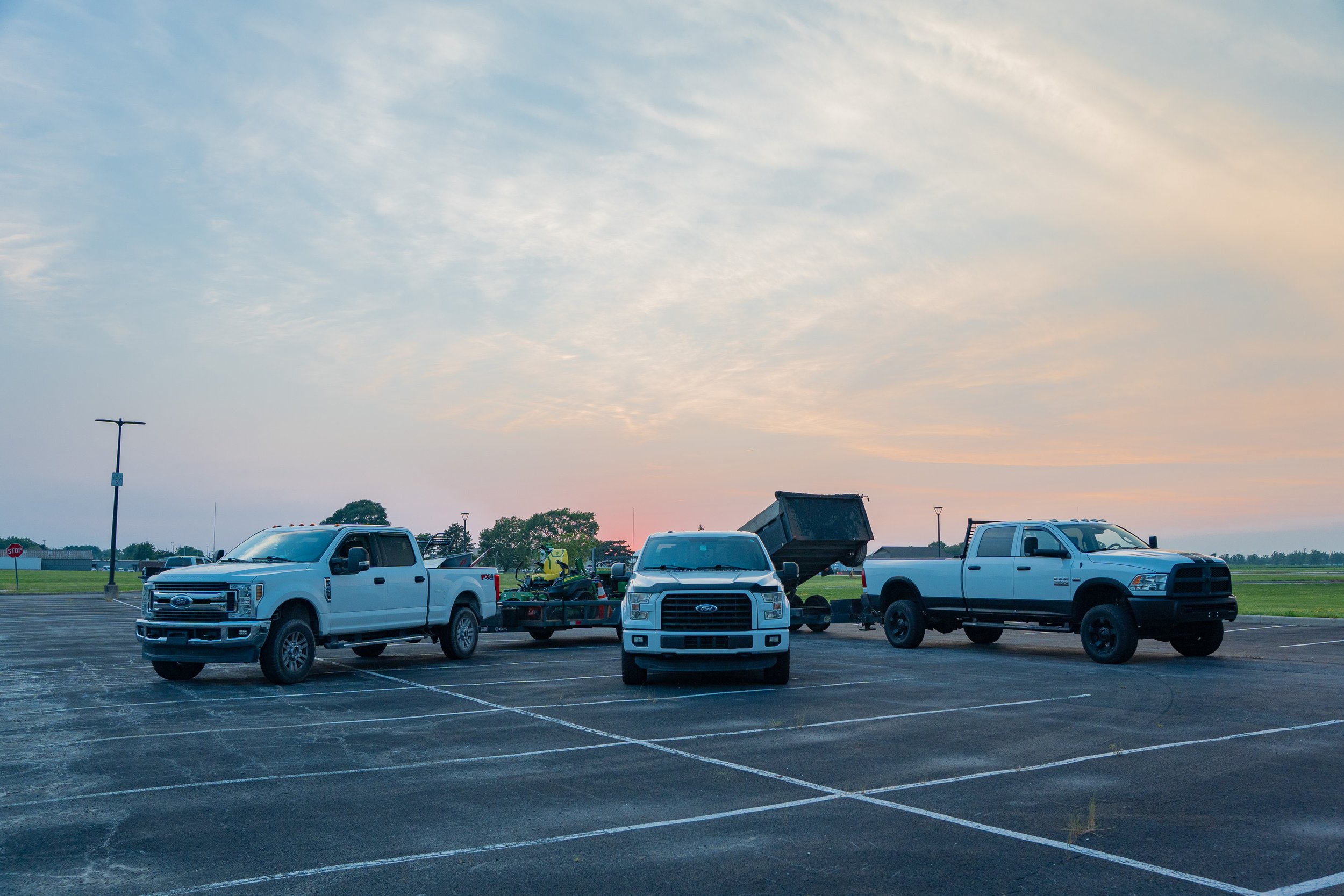 A parking lot at sunset with six trucks parked, some with hauling trailers, and a small equipment vehicle. The sky is partly cloudy with soft pastel colors, and there are a few trees and lampposts in the background.