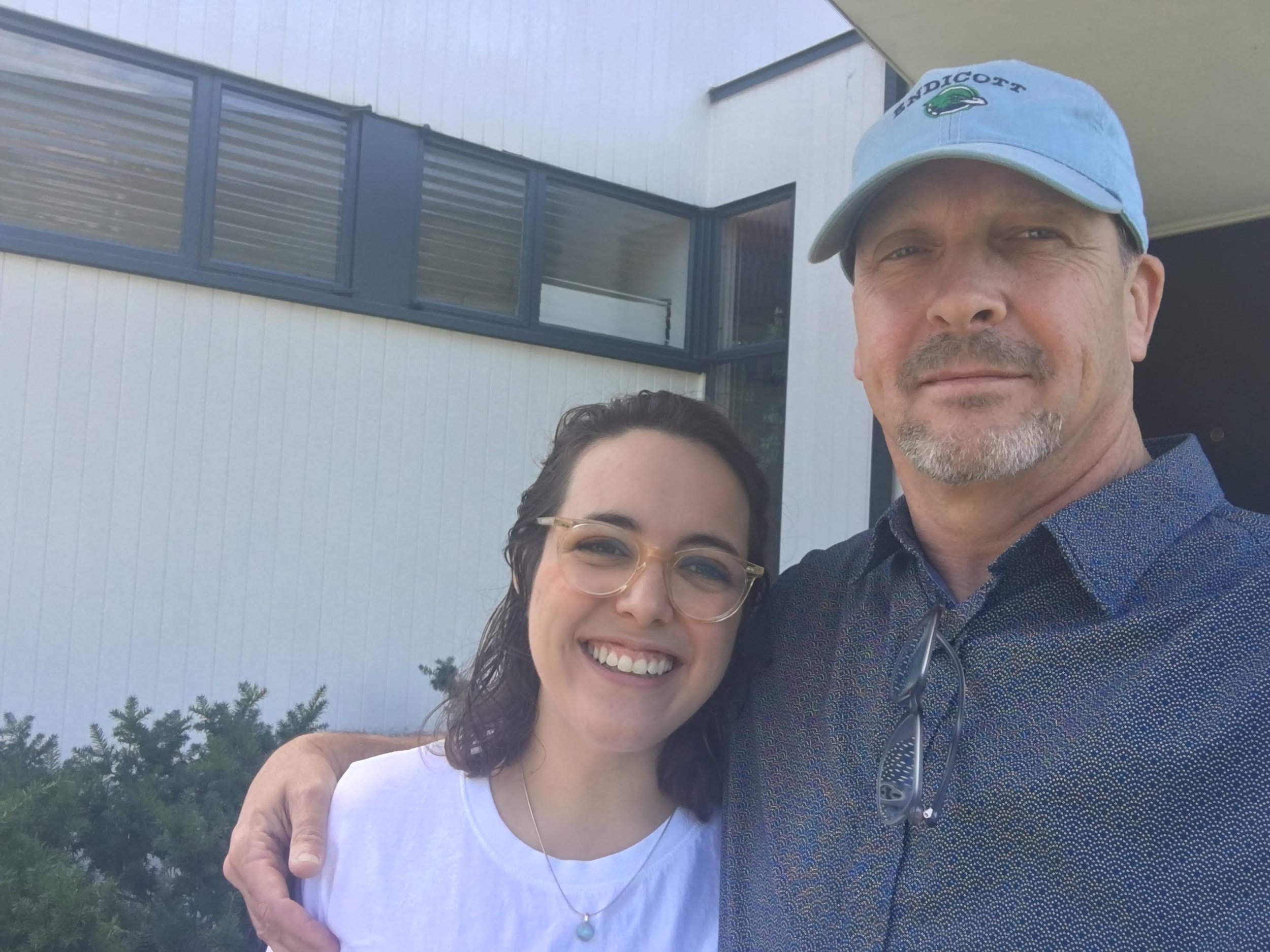 A man and woman taking a selfie outdoors in front of a modern house with large windows and white exterior walls, greenery in the background, the man wearing a light blue hat and a patterned shirt, the woman wearing glasses and a pearl necklace.
