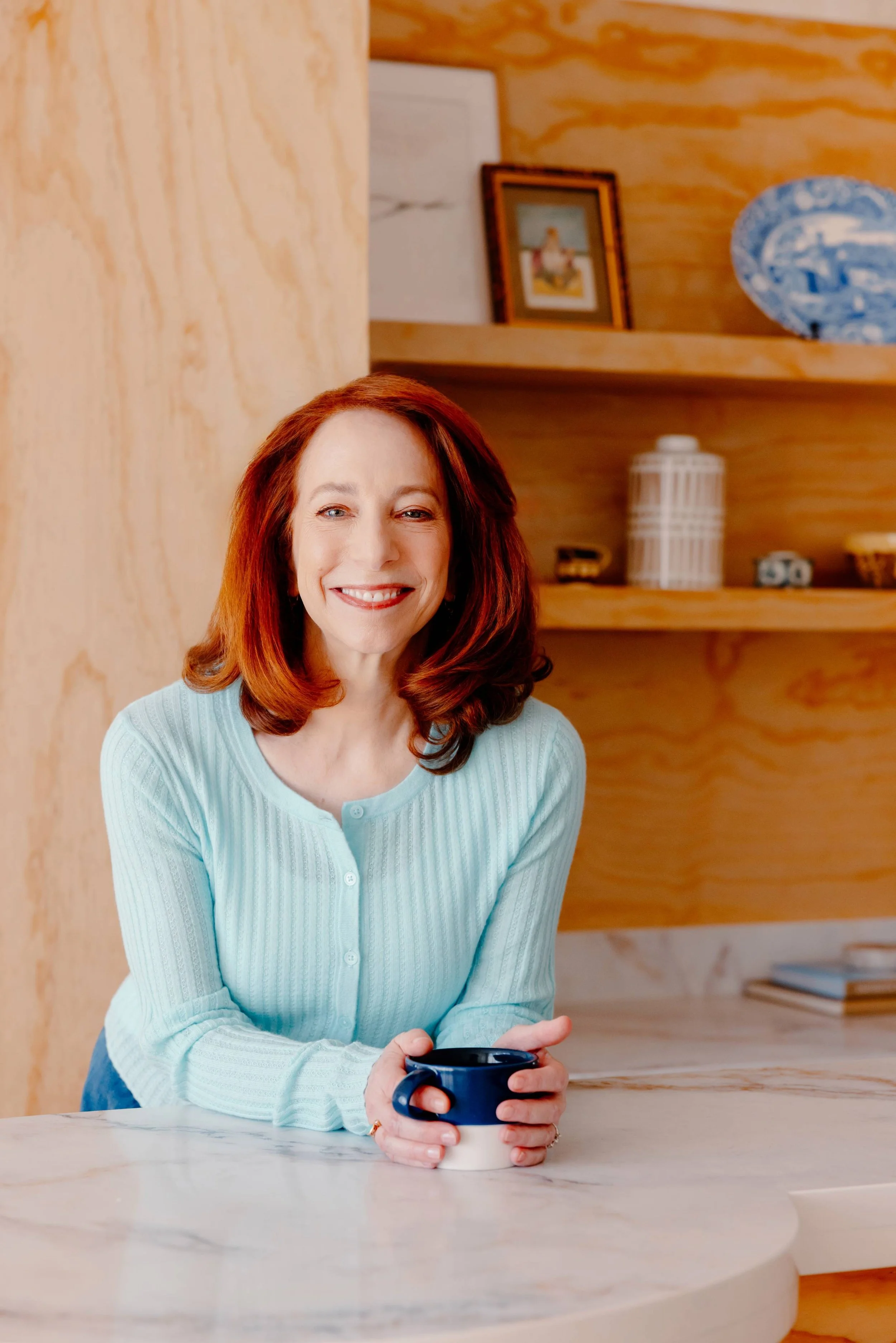 Barbara Rifkind wearing a blue sweater holding a mug and leaning on a table in front of wooden shelves-drop-shadow