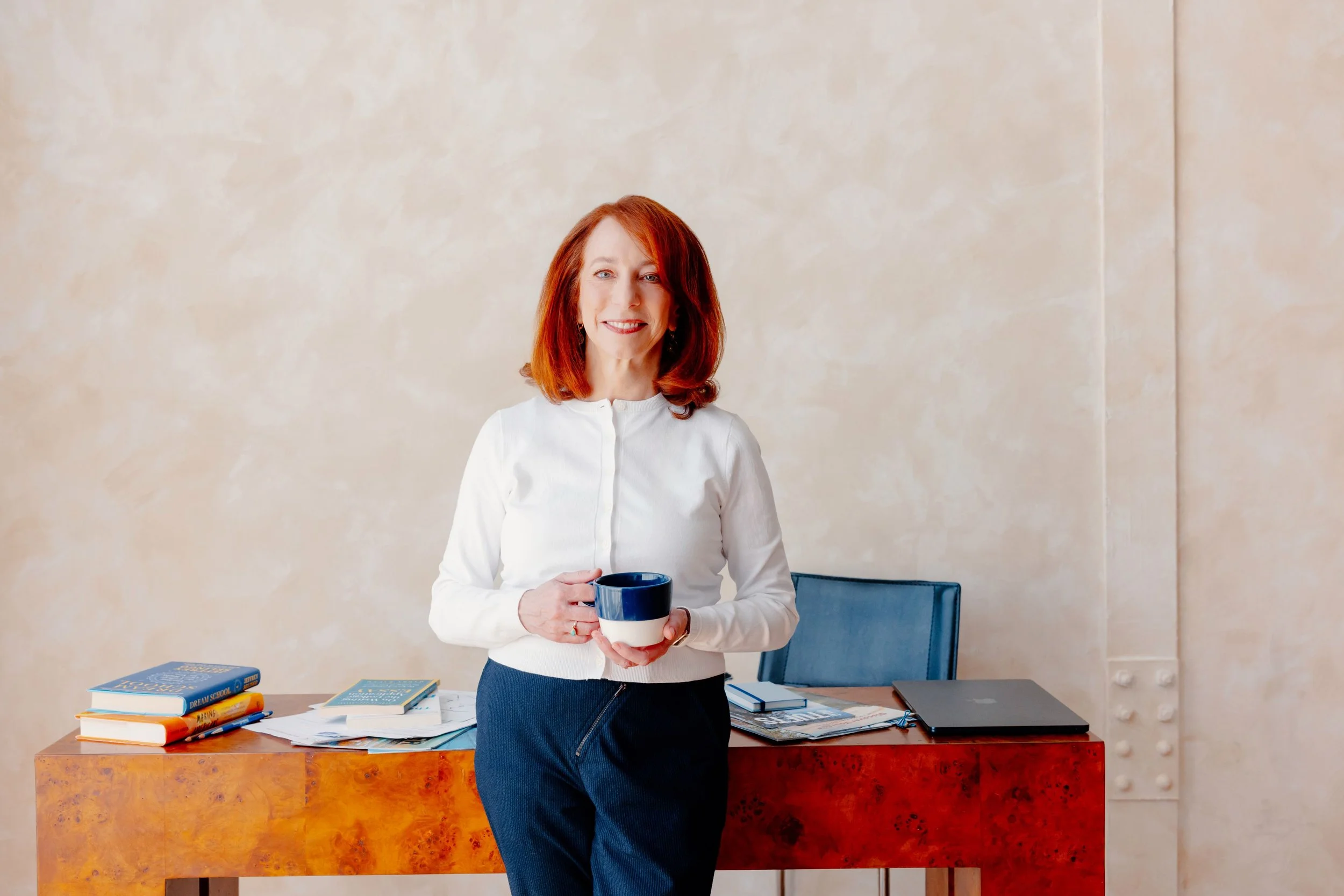 Barbara Rifkind wearing a blue sweater holding a mug and leaning against a large wooden desk-drop-shadow
