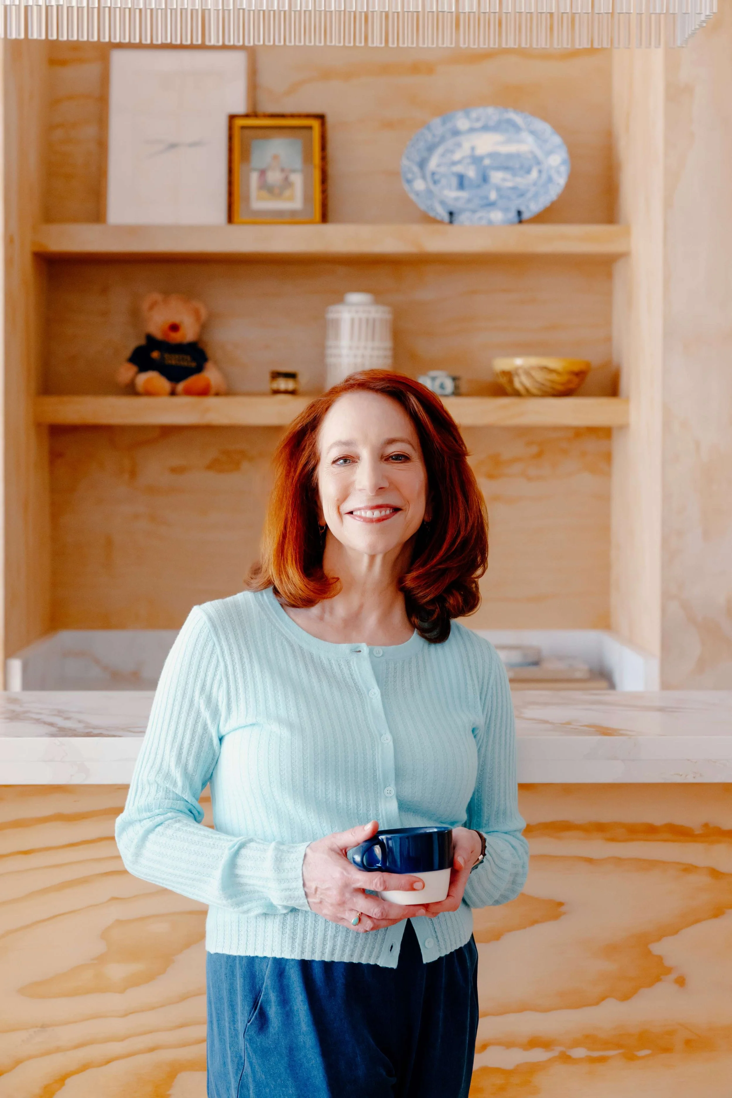 Barbara Rifkind wearing a blue sweater holding a mug and standing in front of wooden shelves-drop-shadow