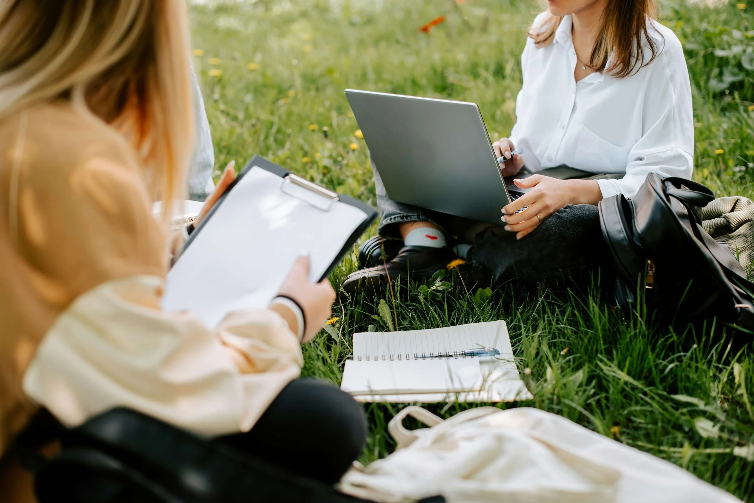 Two teenage girls sitting in the grass with laptops and notebooks-in white card