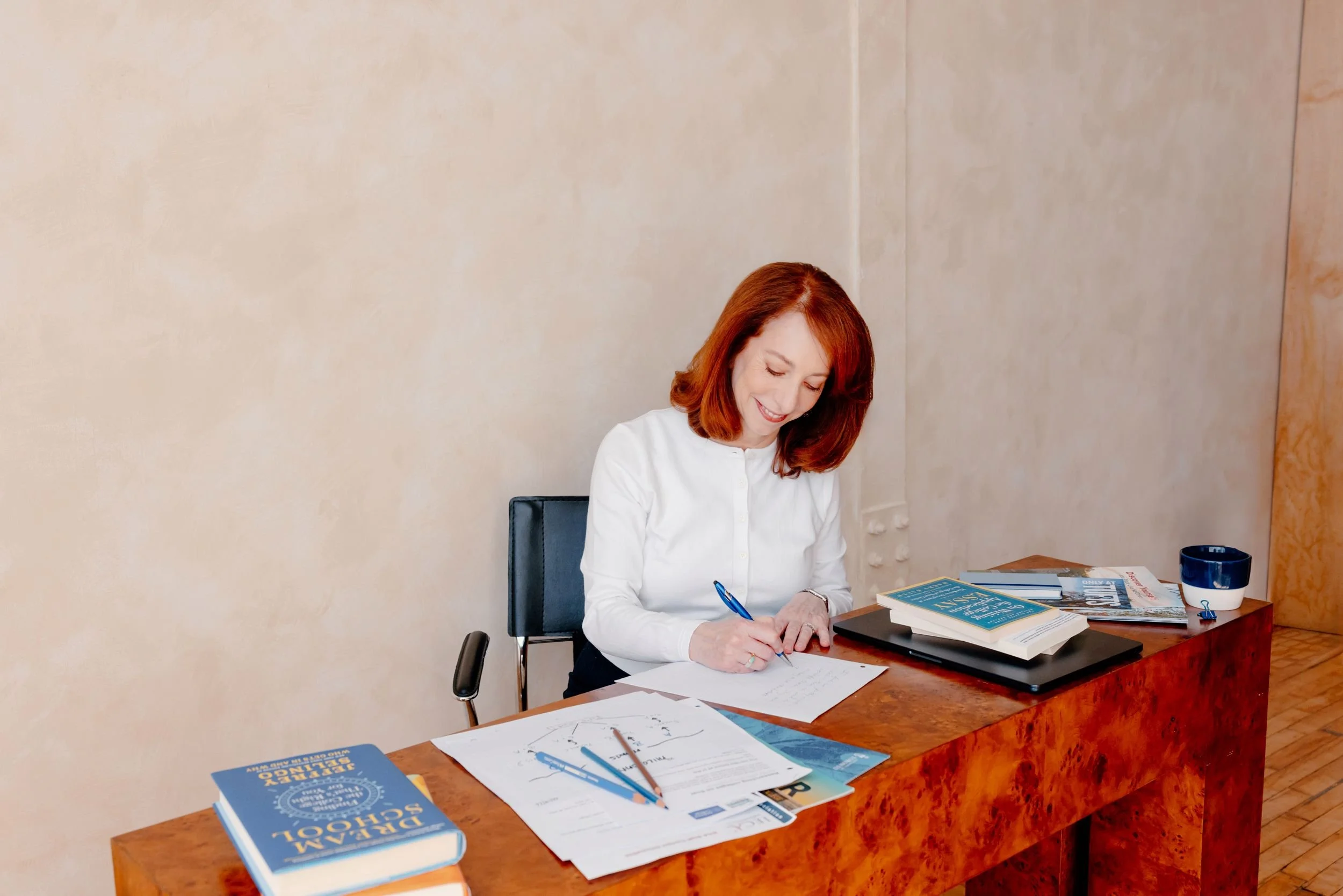 Barbara Rifkind wearing a white sweater sitting at a large wooden desk writing on a piece of paper-in white card