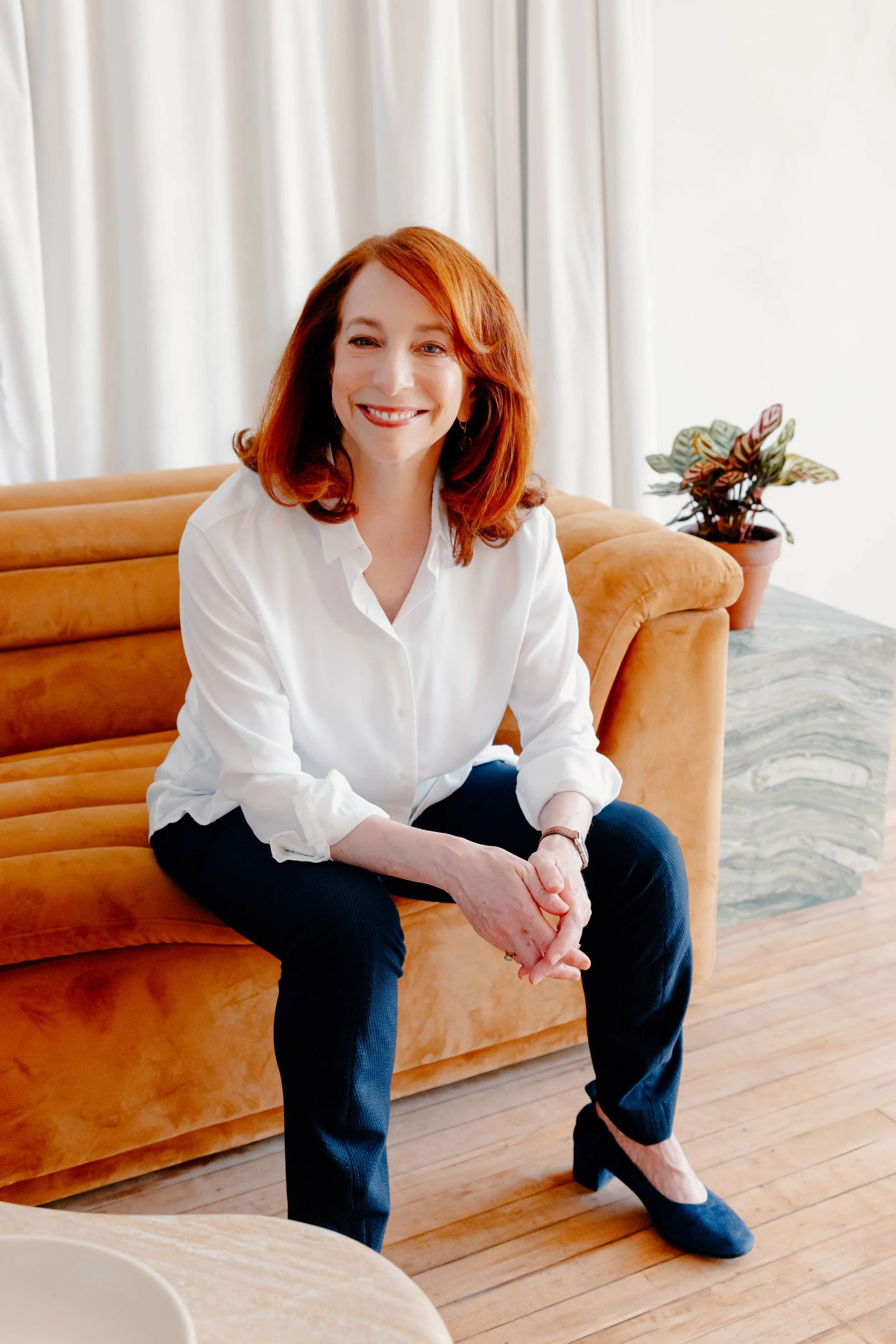 College admissions counselor Barbara Rifkind wearing a white button down and blue pants and sitting on a tan couch-drop-shadow