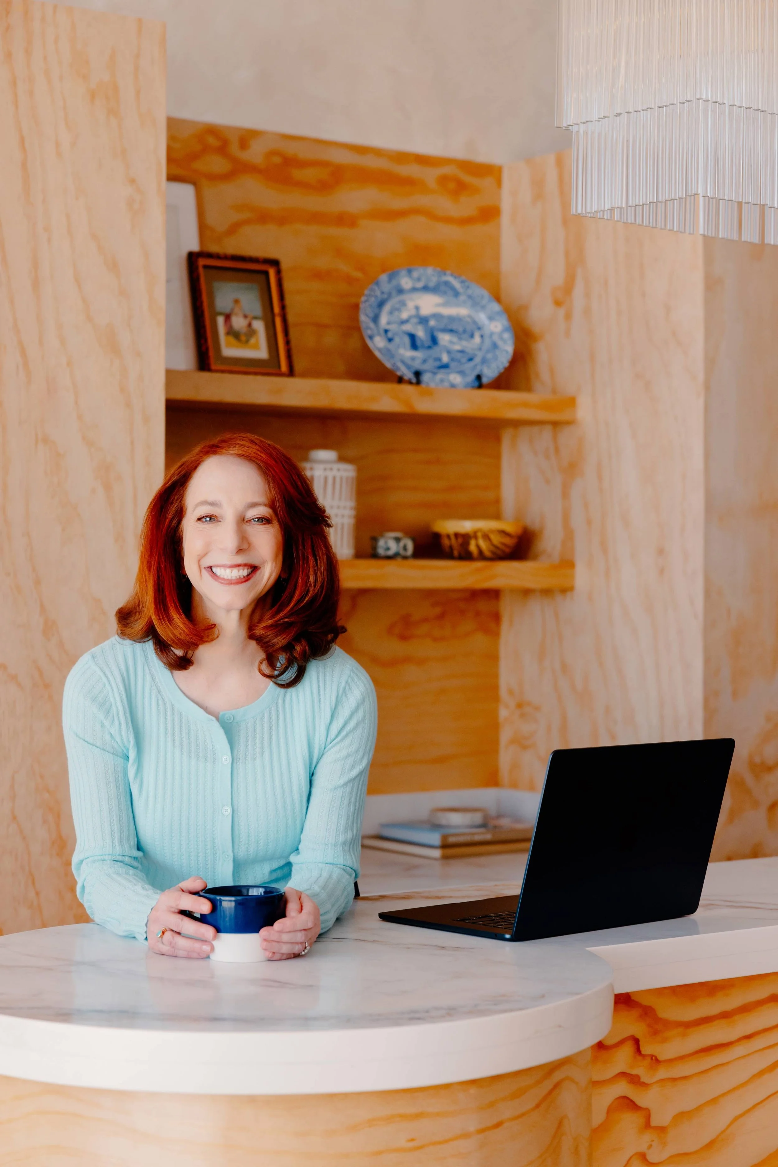 Barbara Rifkind wearing a blue sweater holding a mug and sitting at a table in front of wooden shelves-drop-shadow