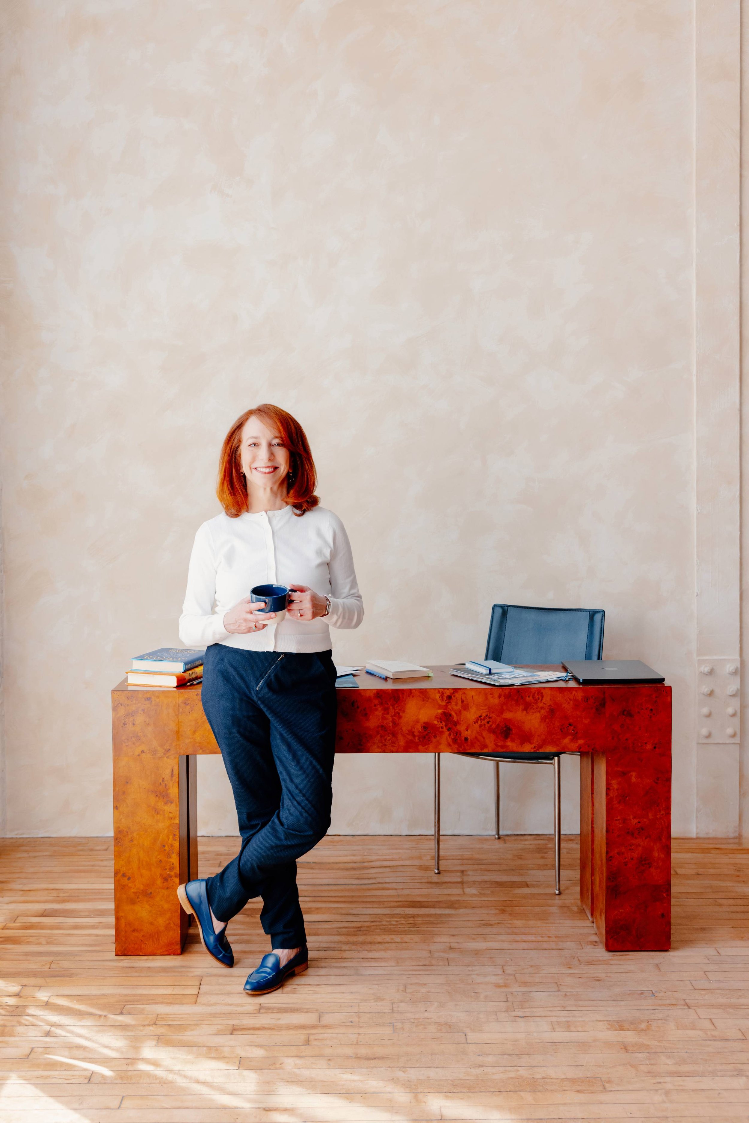 College admissions counselor Barbara Rifkind wearing a white sweater and blue pants leaning against a large wooden desk-drop-shadow