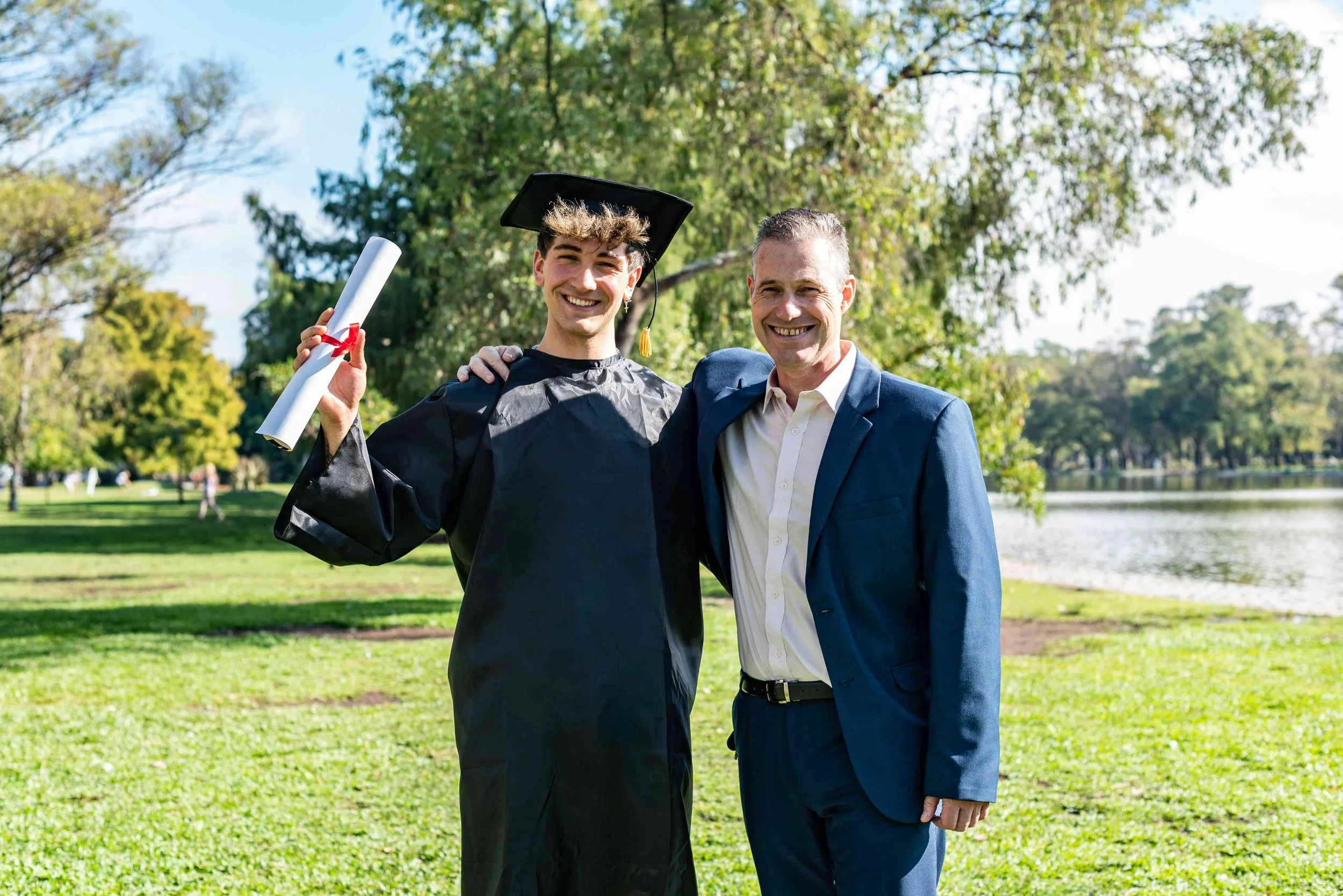 Teenage boy wearing graduation robes and holding a diploma smiling with his father-drop-shadow