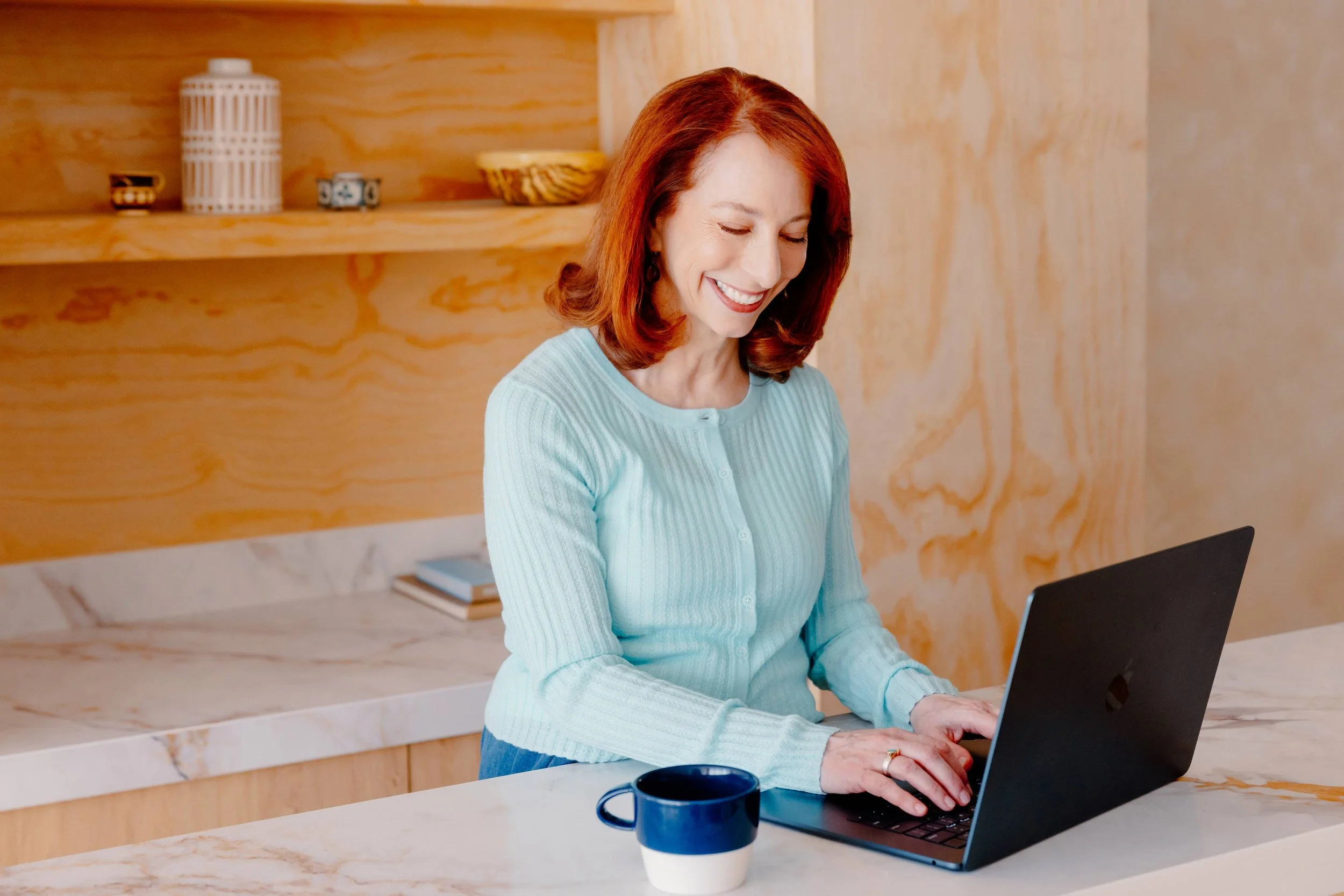 College admissions counselor Barbara Rifkind wearing a blue sweater and typing on a laptop on a table with wooden shelves behind her-drop-shadow