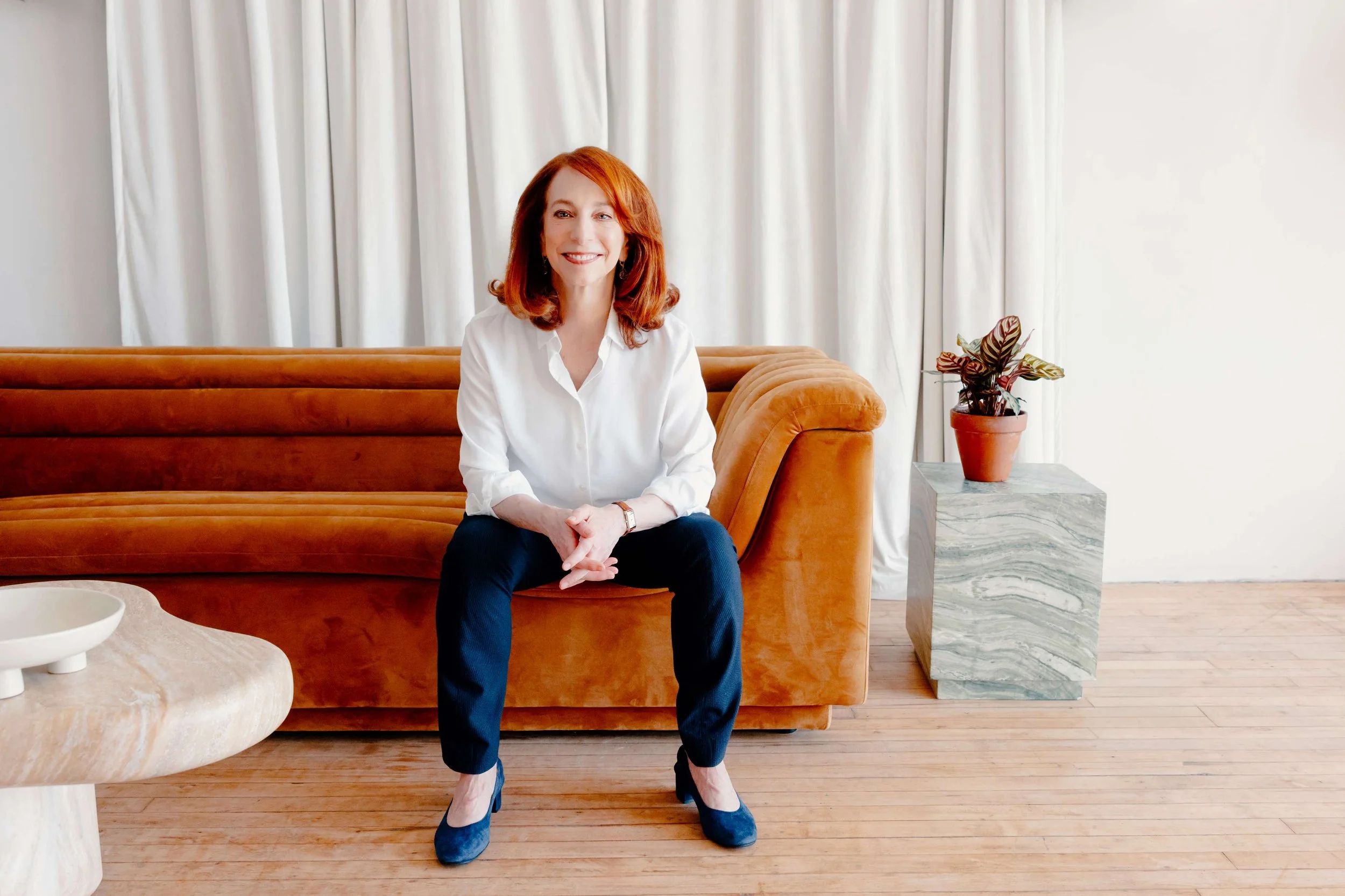 College admissions counselor Barbara Rifkind wearing a white button down and blue pants sitting on a brown velvet couch-drop-shadow