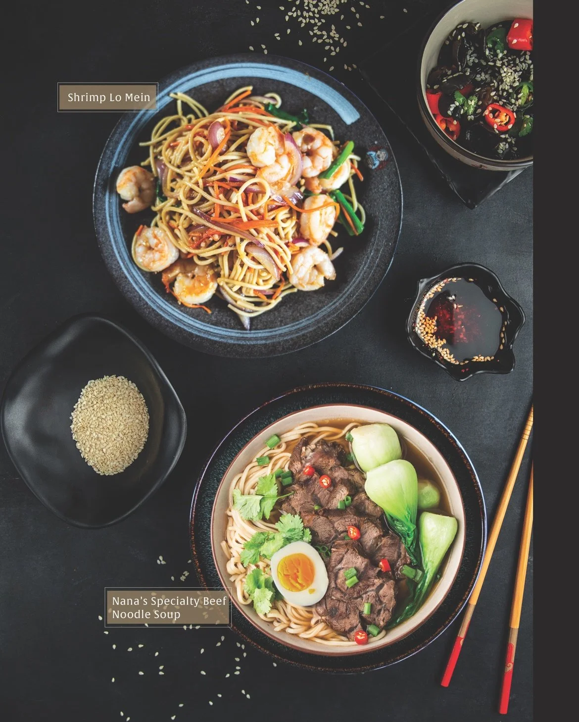 A table with a bowl of shrimp lo mein, a bowl of beef noodle soup with half a boiled egg, a small dish of sesame seeds, a small dish of soy sauce with sesame seeds, a bowl of seaweed salad with sesame seeds and chili peppers, and chopsticks.