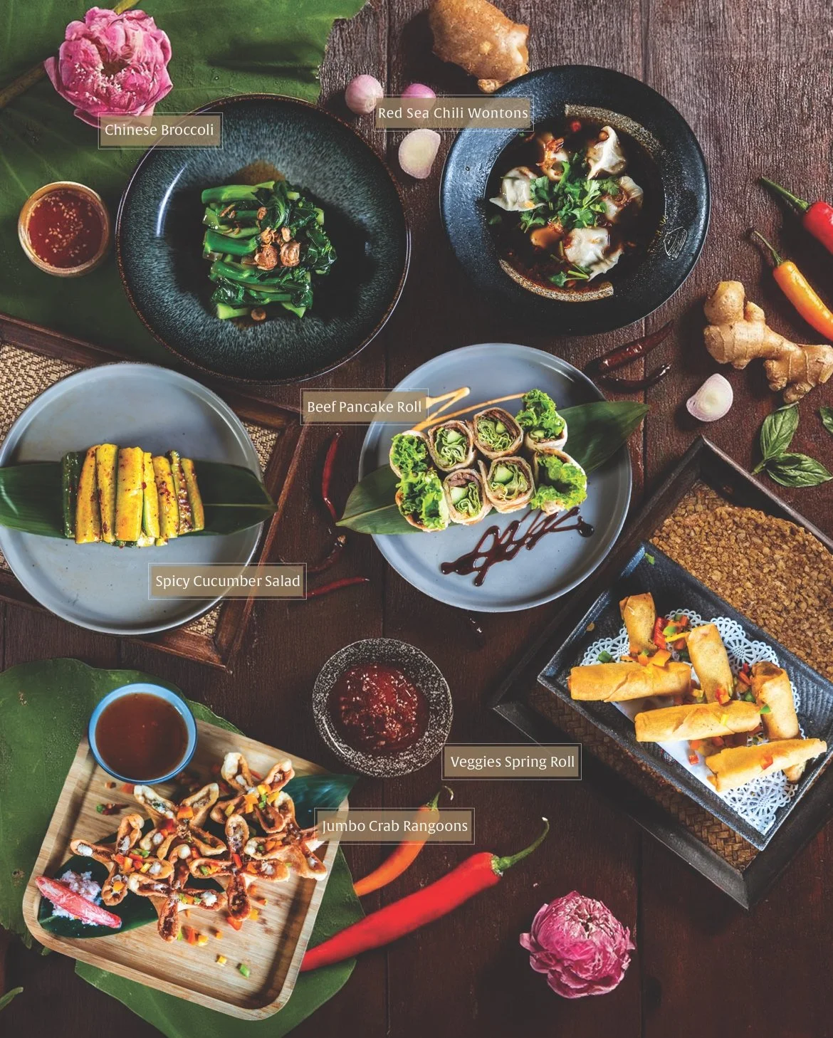 A variety of Asian dishes arranged on a wooden table, including Chinese broccoli, red sea chili wontons, beef pancake roll, spicy cucumber salad, jumbo crab rangoons, and veggie spring rolls, with small bowls of sauces and decorative flowers.