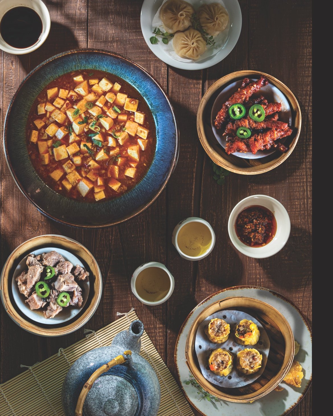 An assortment of Asian dishes on a wooden table, including a large bowl of mapo tofu, a plate of dumplings, a basket of fried chicken strips with jalap CPef slices, steamed dim sum, a bowl of dipping sauce, and two cups of tea.