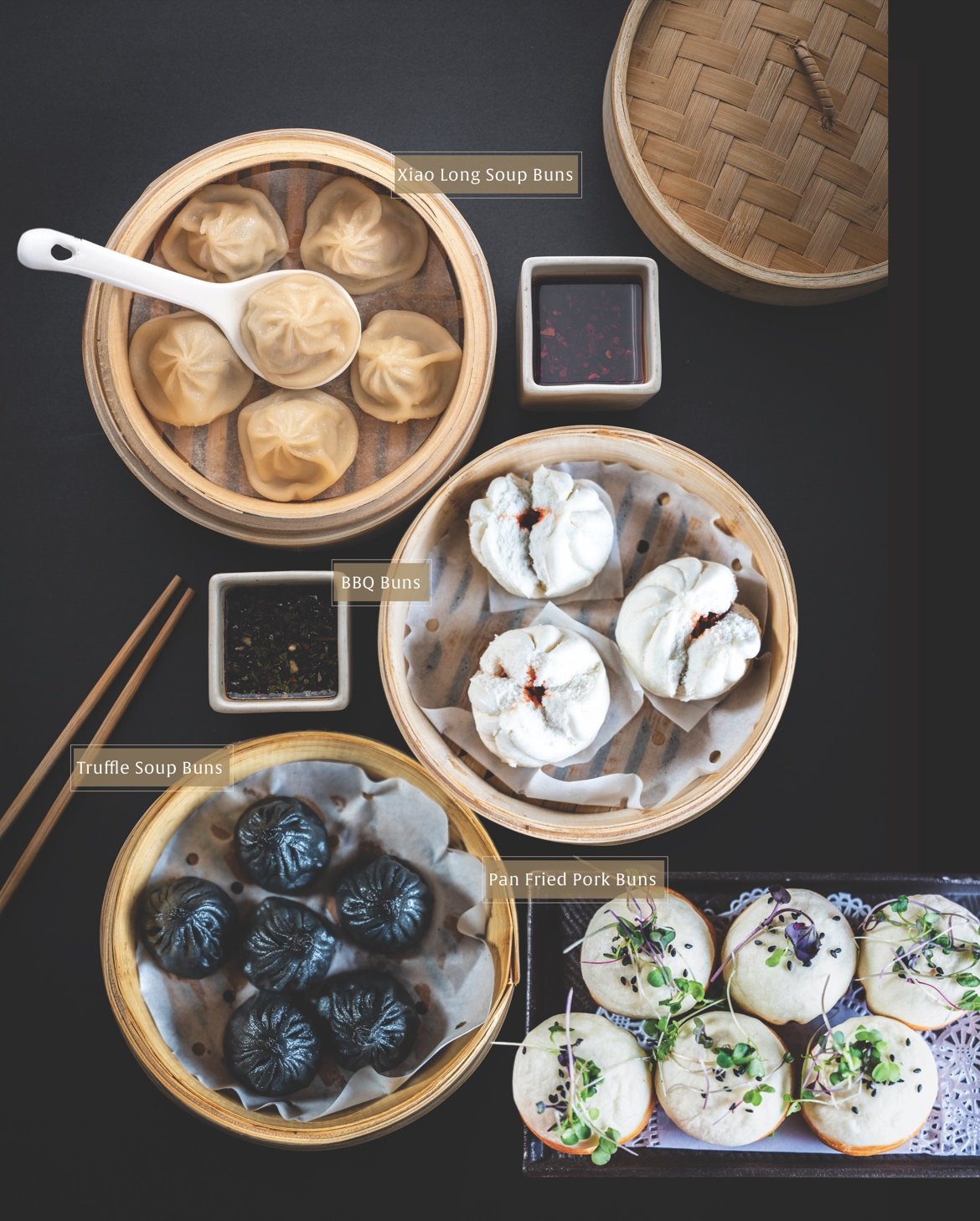 Assorted steamed buns on a table, including Xiao Long Soup Buns, BBQ Buns, Truffle Soup Buns, Pan Fried Pork Buns, with dipping sauces and chopsticks.