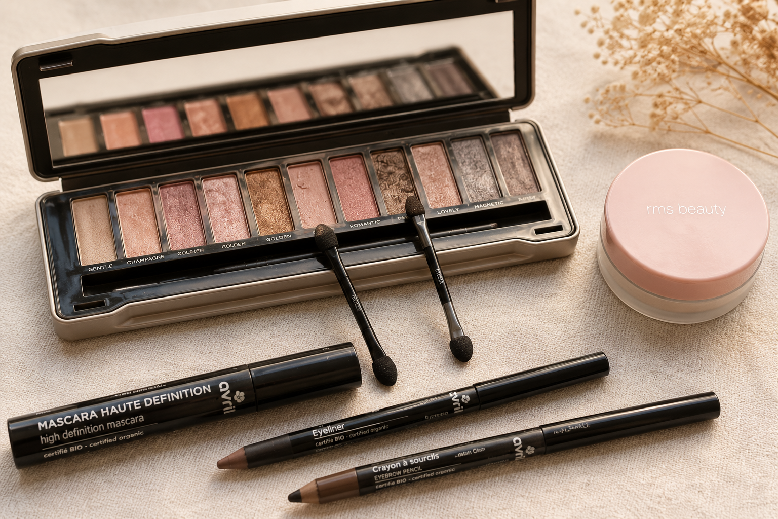 Une palette de fards à paupières avec un miroir, deux pinceaux, un mascara, un eyeliner, un crayon à sourcils et une boîte de crème de beauté sur une surface beige.