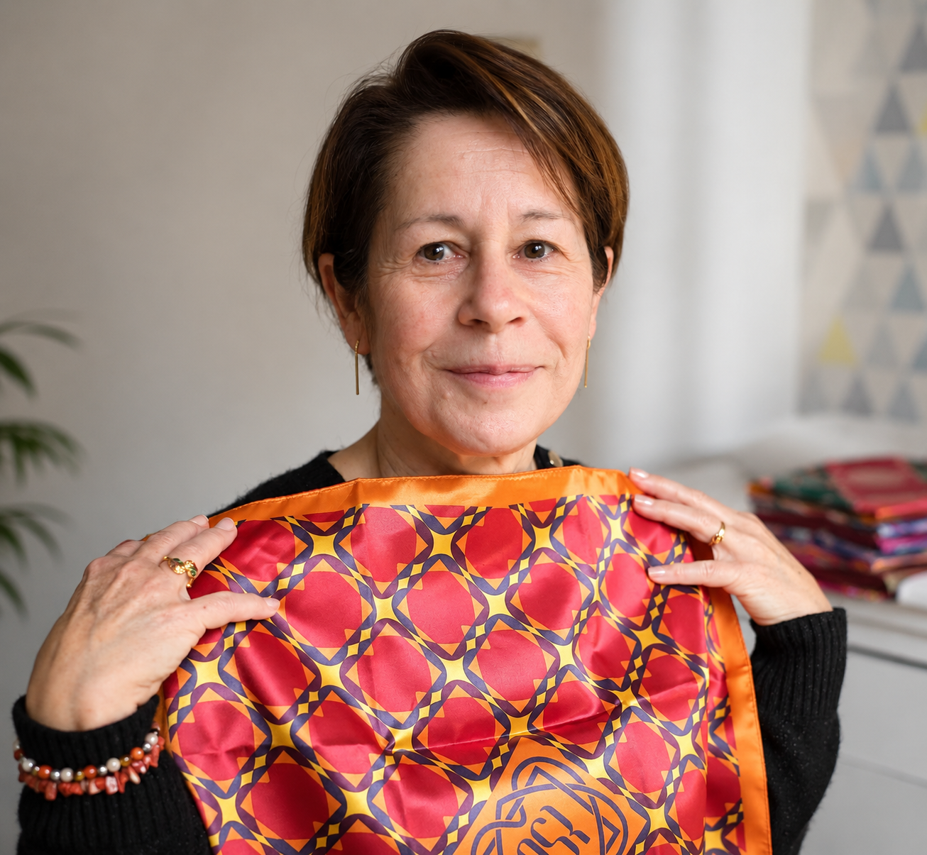 Une femme d'âge moyen tient un foulard coloré rouge et orange avec un motif géométrique, elle sourit doucement dans un intérieur lumineux.