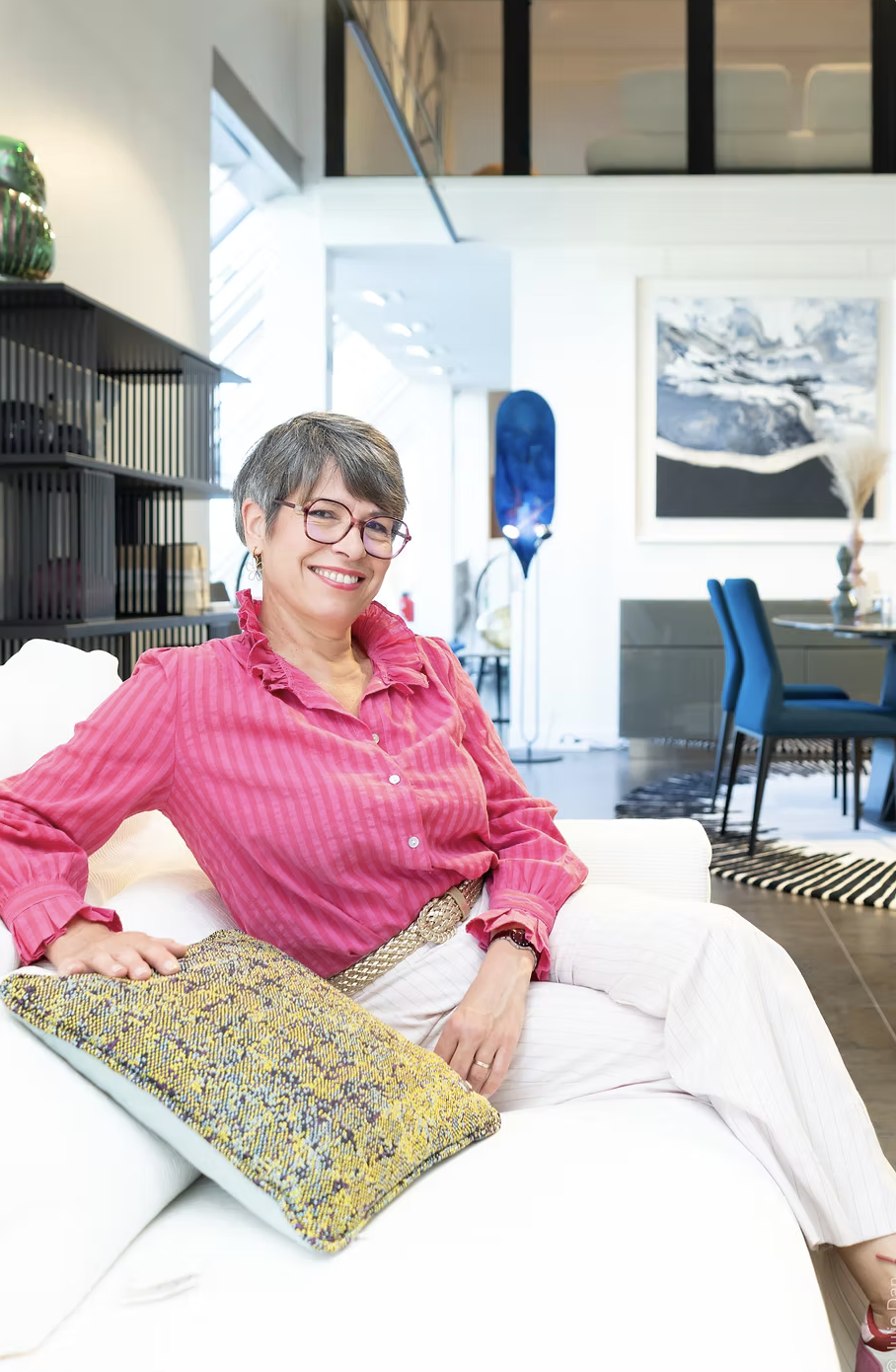 Une femme souriante portant des lunettes et une chemise rose à rayures, assise sur un canapé blanc dans un intérieur moderne et lumineux avec des œuvres d'art et des meubles bleus en arrière-plan.