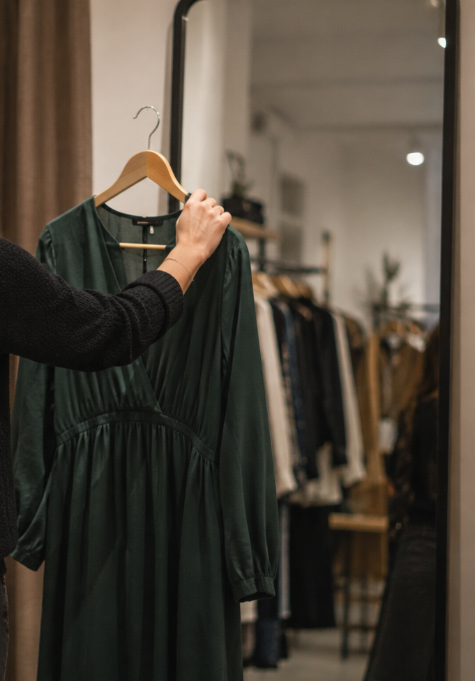 Une personne tient une robe verte sur un portant dans un magasin de vêtements, vue partielle du bras et de la main.