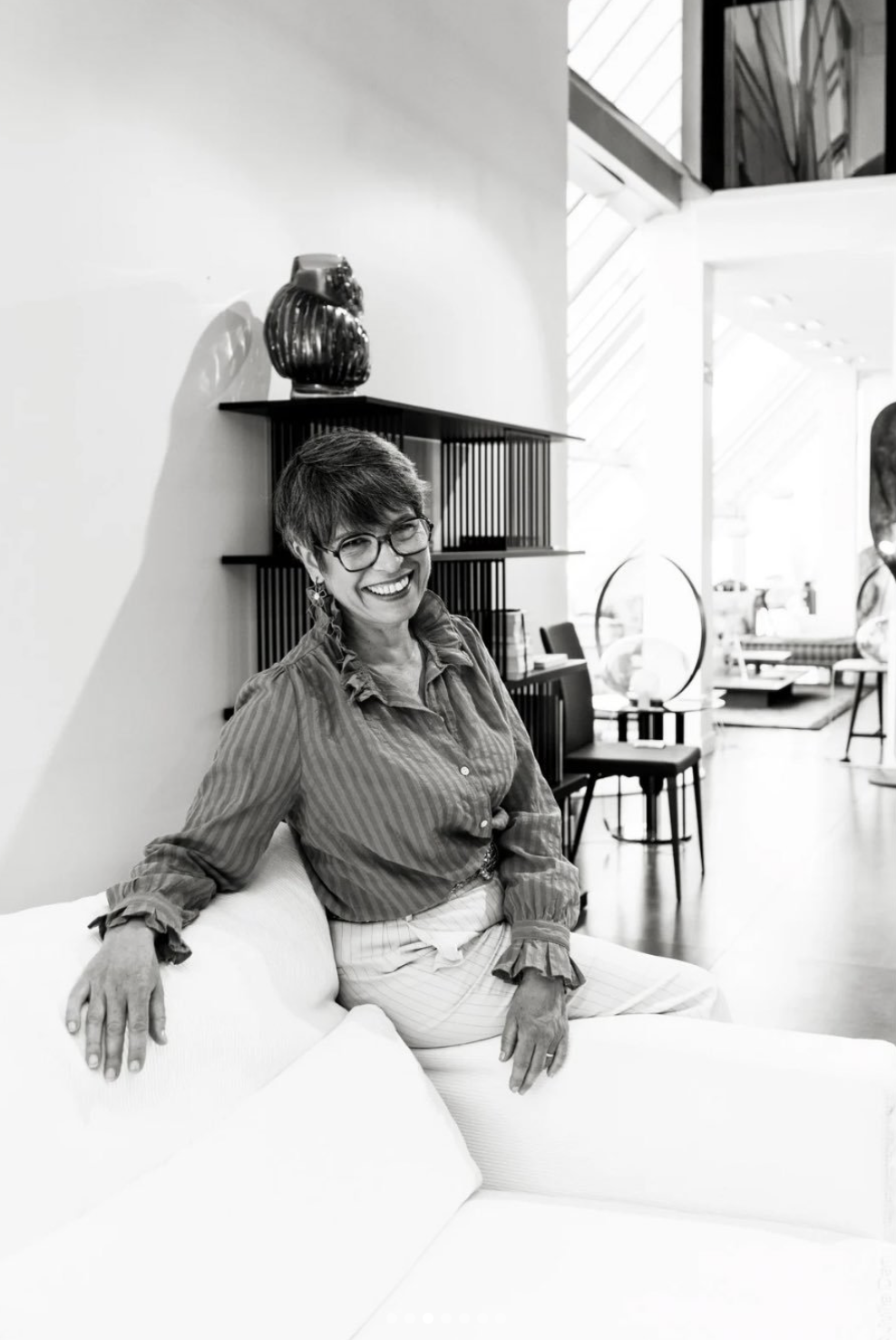 Une femme souriante avec des lunettes assise sur un canapé blanc dans un intérieur moderne.