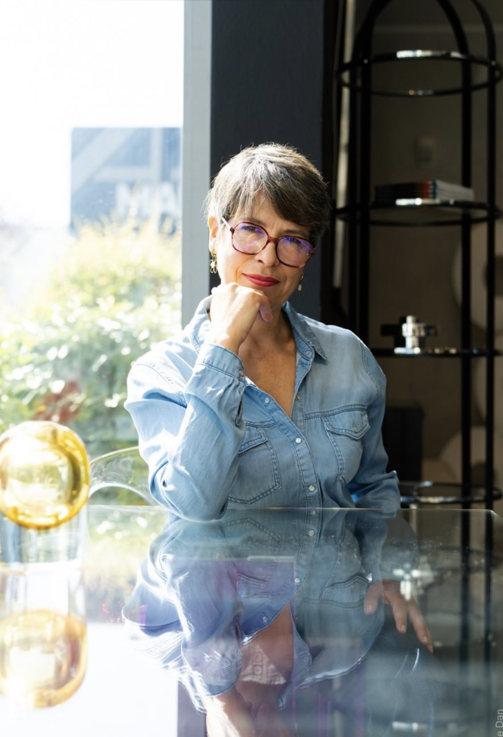 Femme souriante avec lunettes assise à une table en verre près d'une fenêtre, avec une bouteille de vin blanc ou d'eau et un citron dans un verre. Décor intérieur moderne.