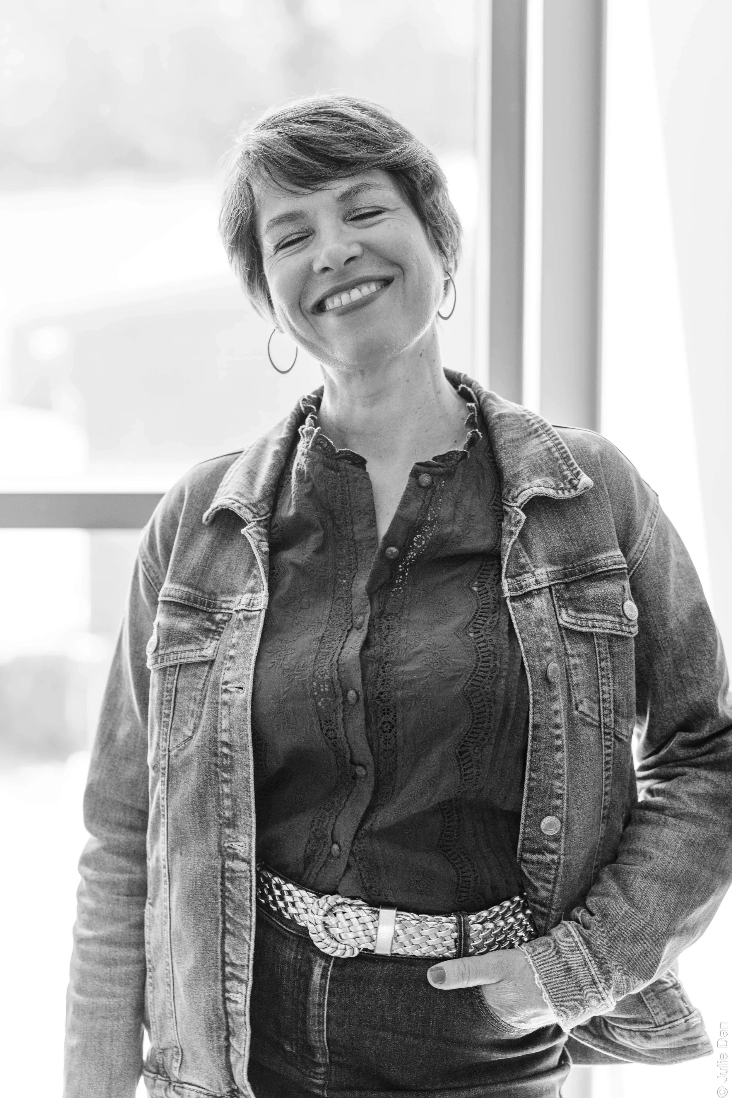 Portrait en noir et blanc d'une femme souriante portant une veste en jean, un haut texturé, une ceinture tressée, et des boucles d'oreilles, debout près d'une fenêtre.