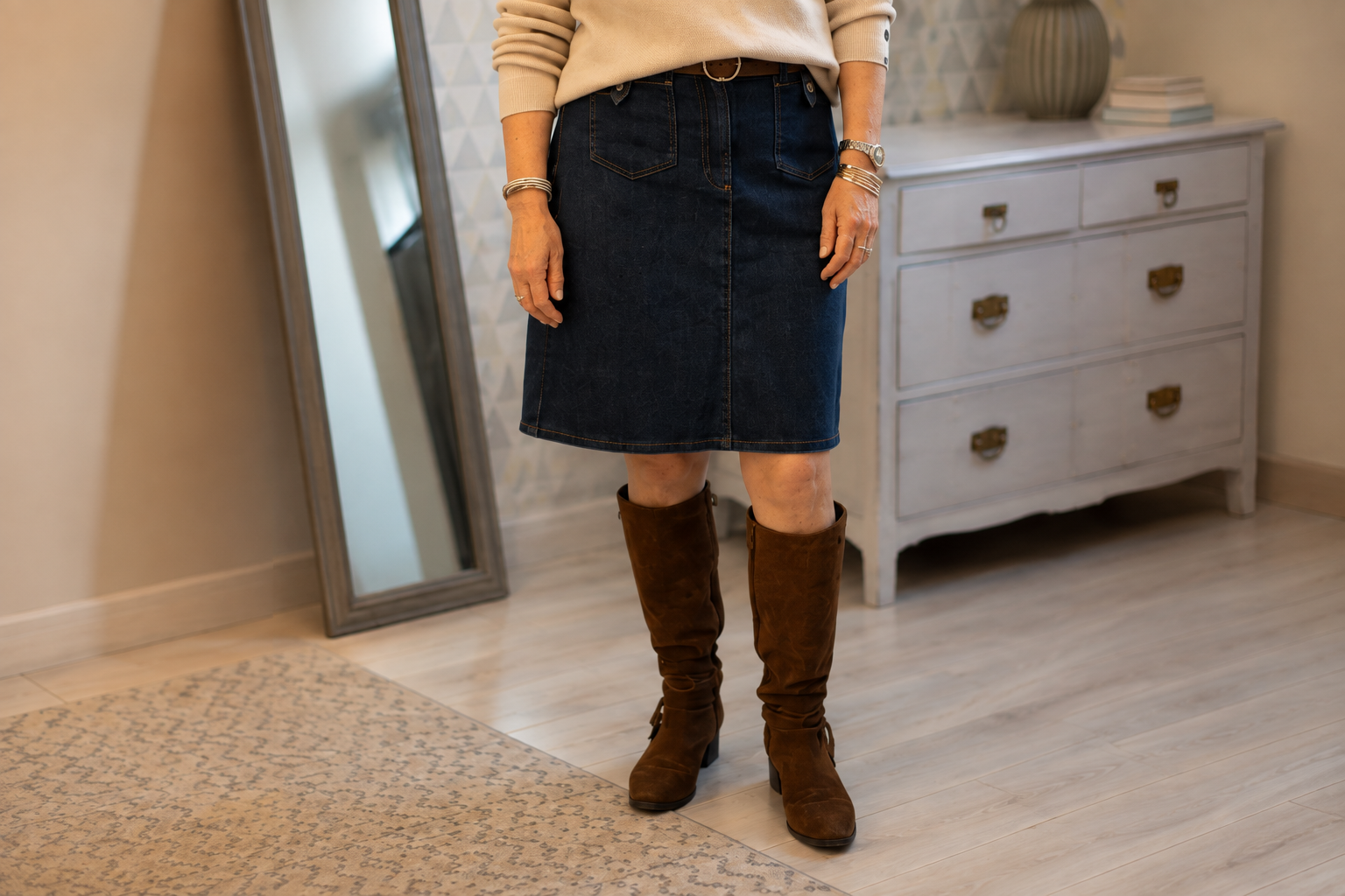 Une personne portant une jupe en denim marron clair, des bottes en cuir marron foncé, un bracelet et une montre, dans une pièce avec un miroir et une commode blanche.