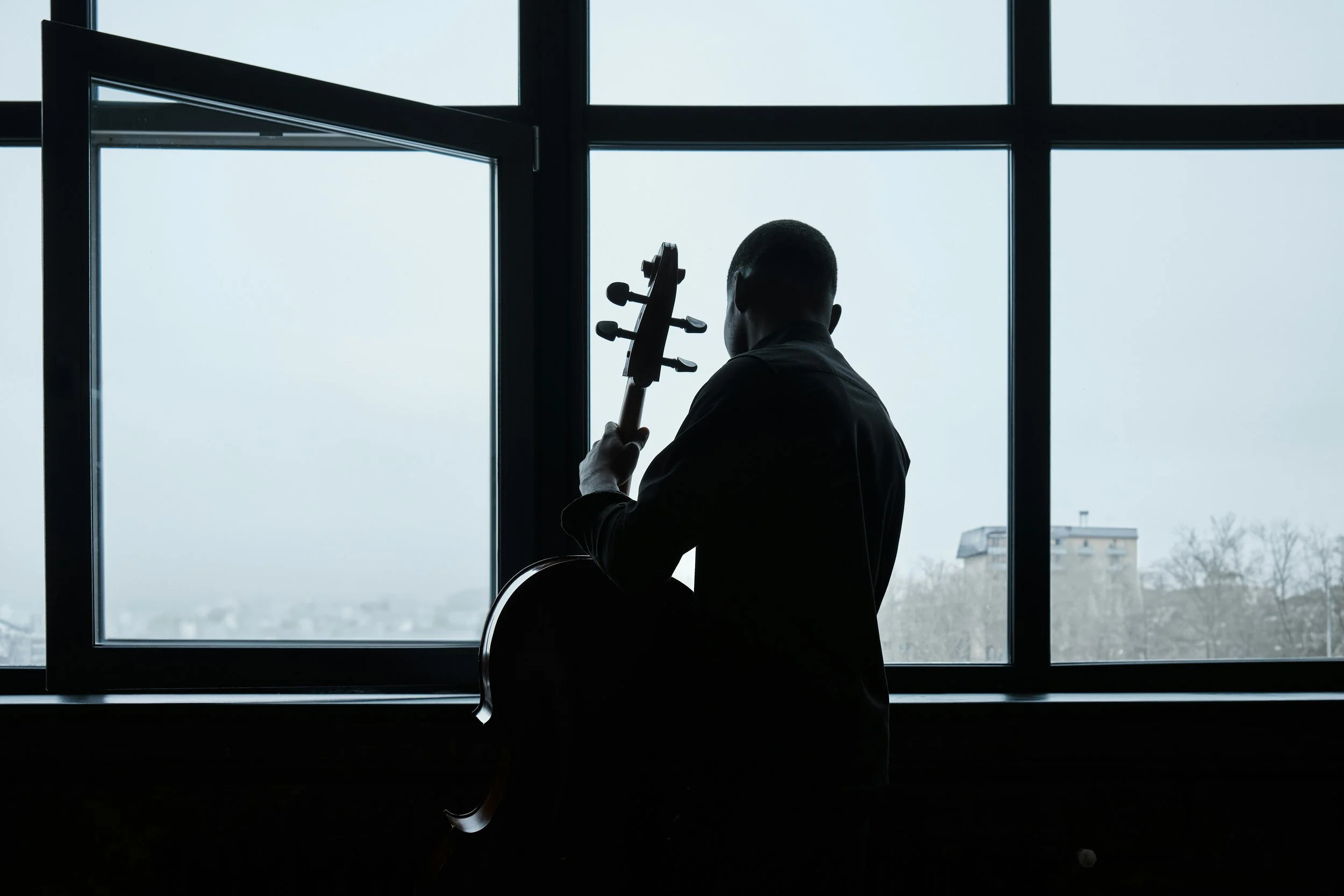 Silhouette of a person holding a guitar, sitting in front of large window with city view, during daytime.