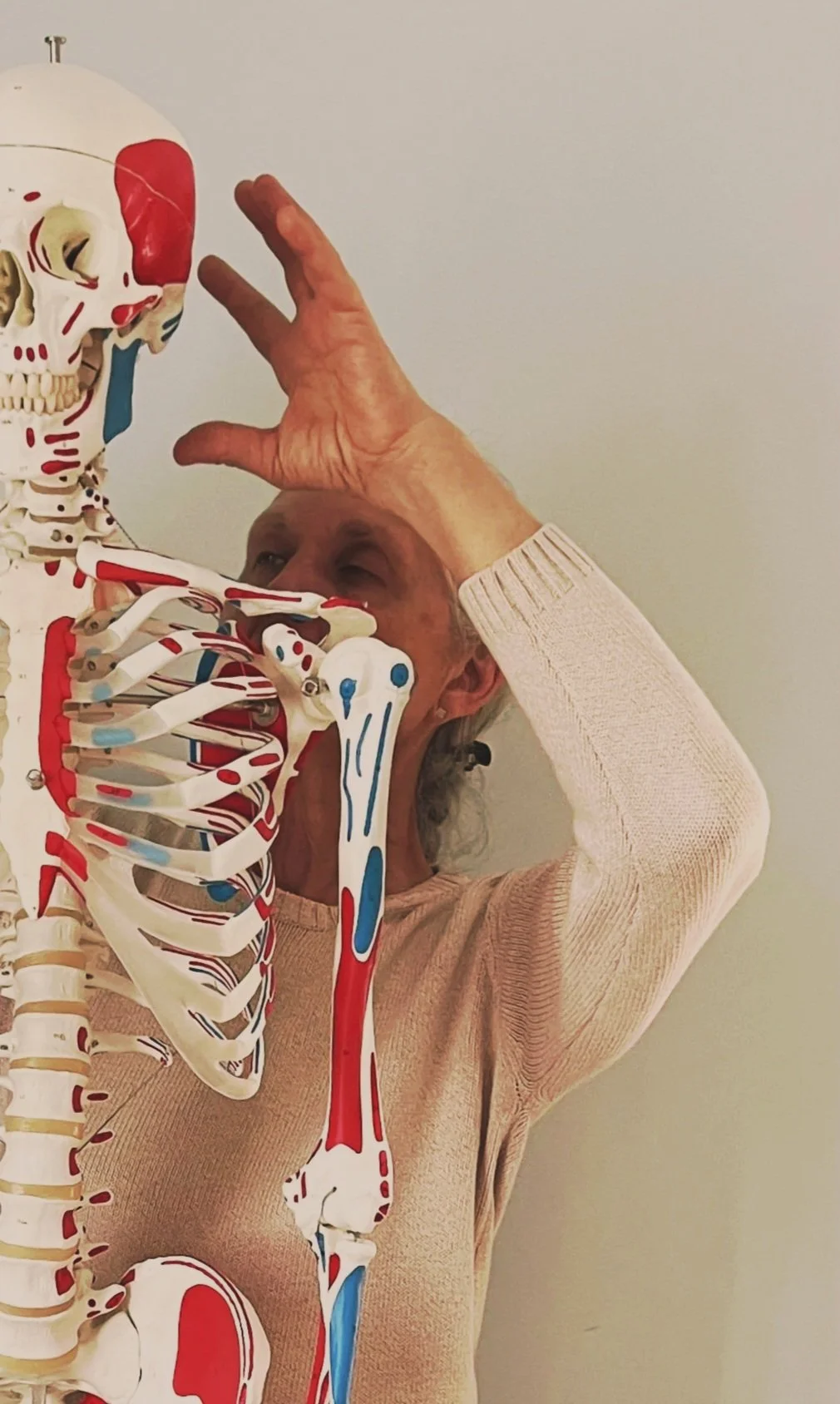 A woman interacts with a human skeleton model, gently touching its skull with her hand. The woman is wearing a beige sweater and is focused on the skeleton.