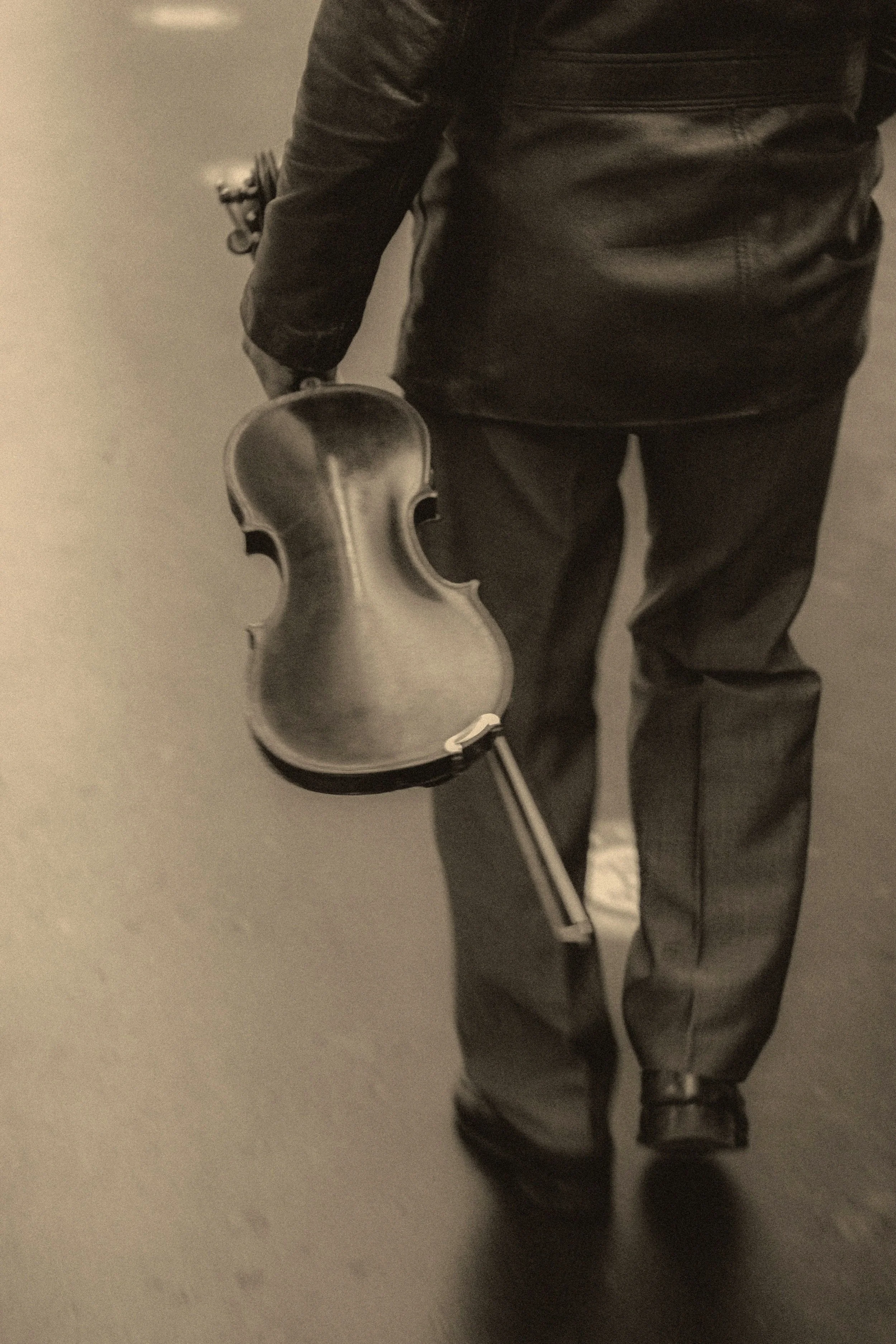 A person dressed in black leather jacket and dark pants holding a violin, standing on a beige floor.