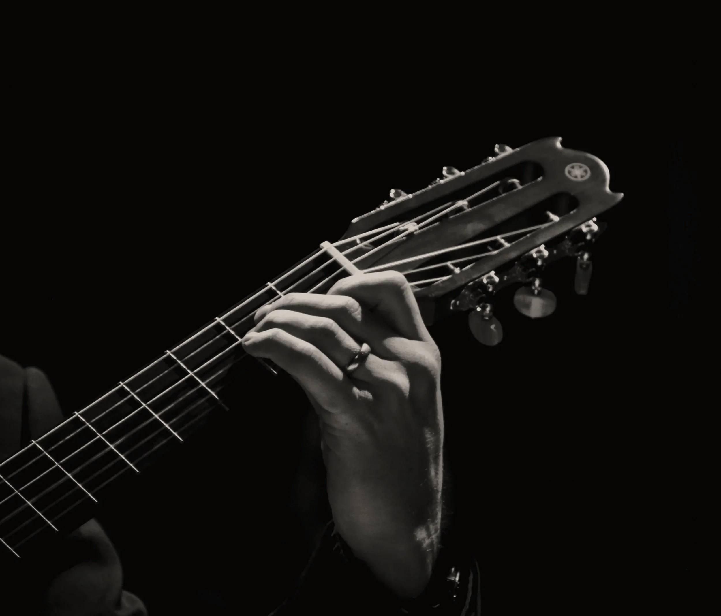 A black and white image showing a hand playing a guitar, focusing on the headstock and tuning pegs against a dark background.