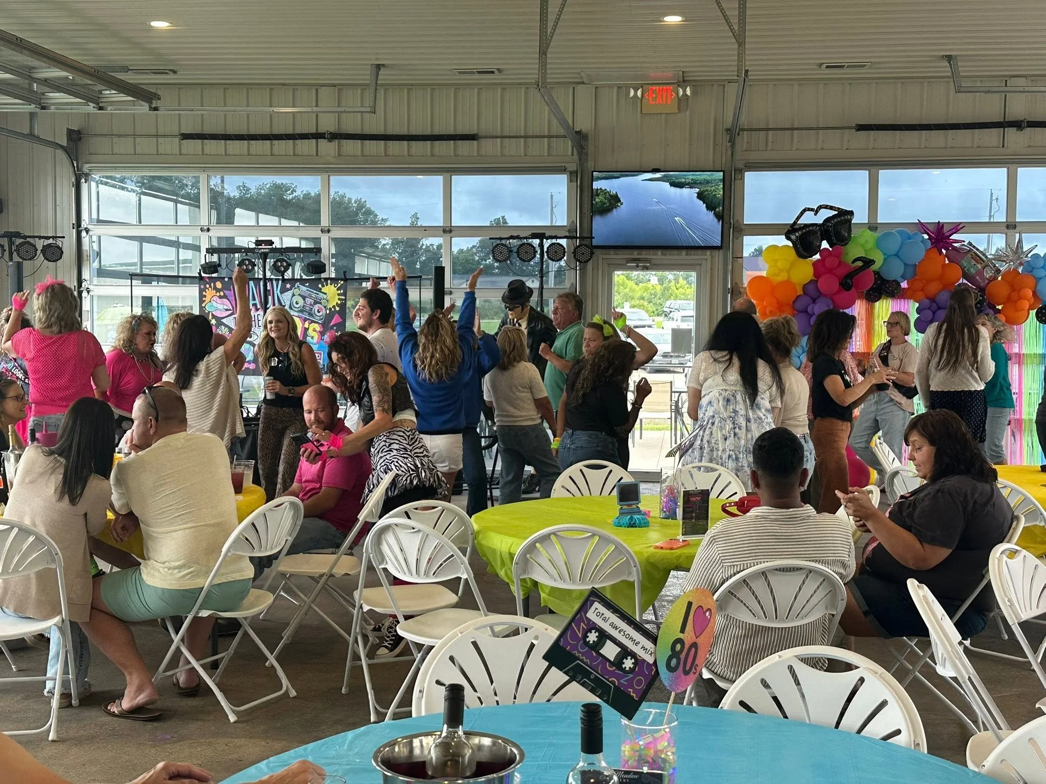 Indoor party scene with people dancing and socializing under a high ceiling with large windows showing sky and trees outside. Colorful balloons and decorations adorn the walls. Tables with yellow and blue tablecloths and drinks are visible in the for