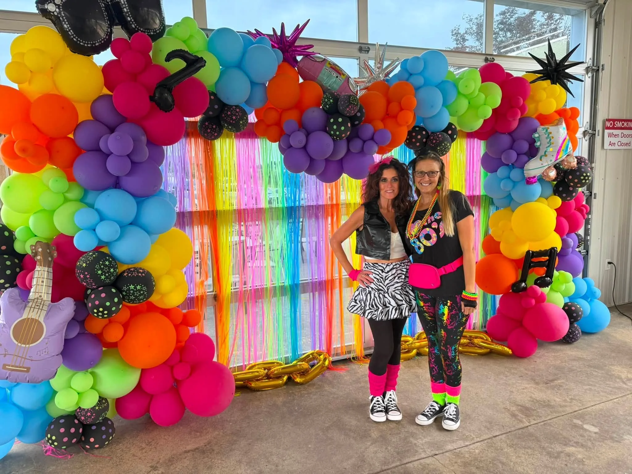 Two women standing in front of a colorful balloon arch with rainbow fringe backdrop, dressed in bright, playful outfits, celebrating a festive event.