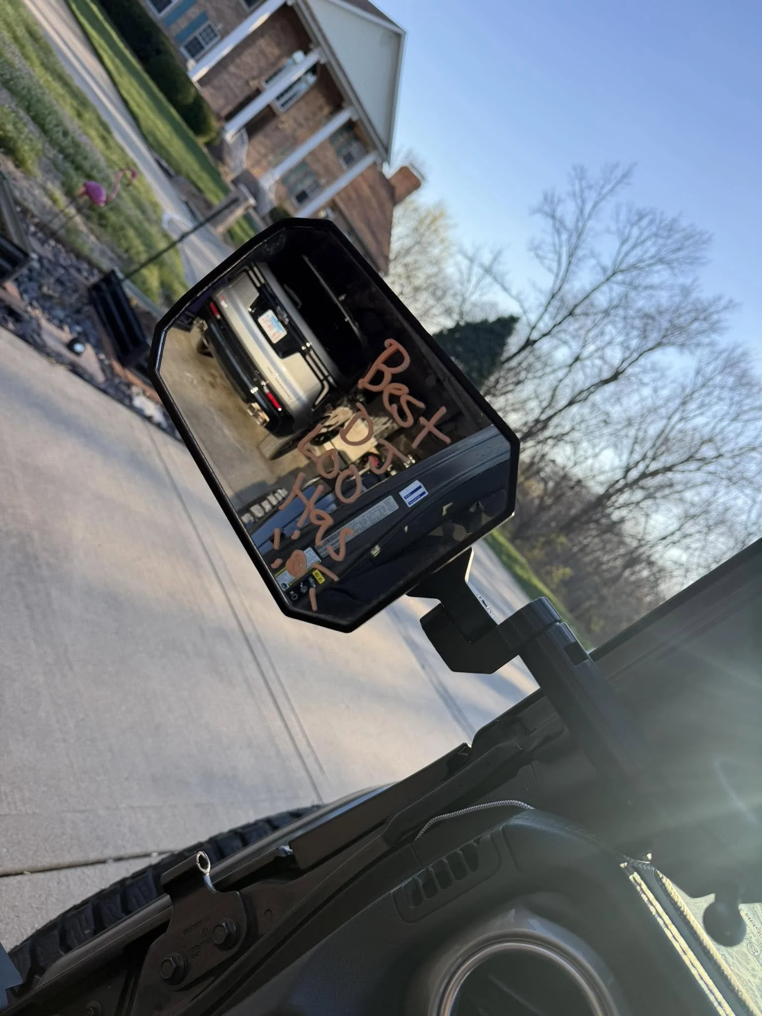A car's side mirror reflects a parked vehicle with a message written in red on the mirror indicating a reminder to rest, with a residential house and trees visible in the background.