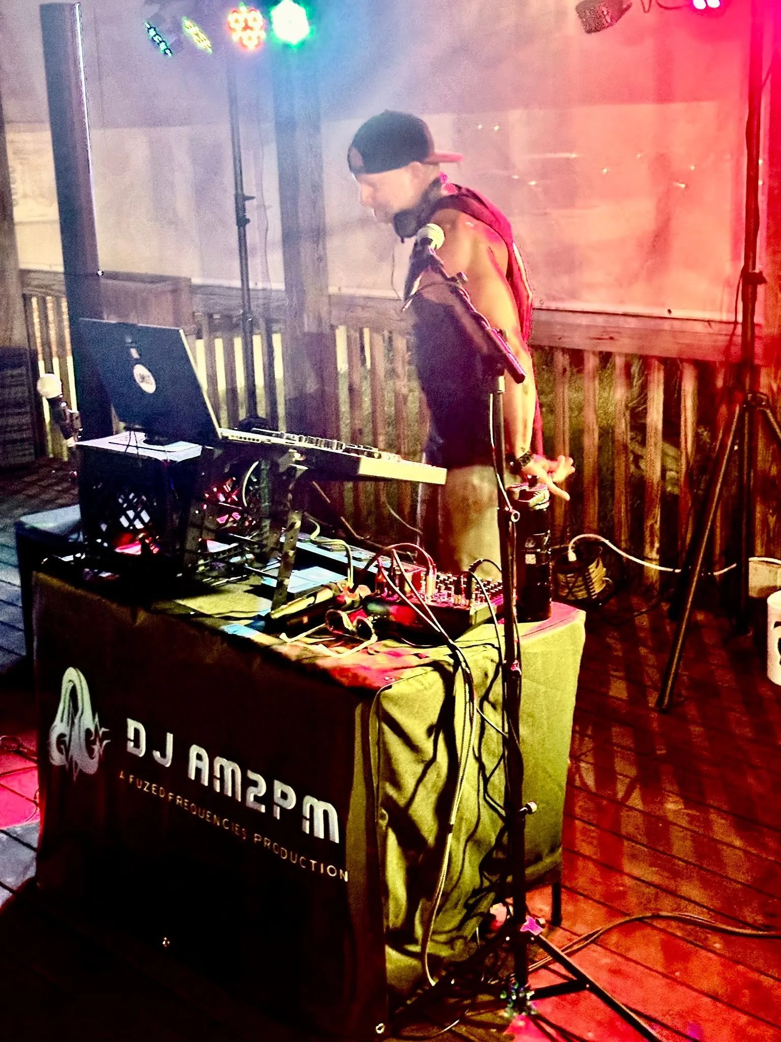A DJ stands behind a table with music equipment, wearing a dark hoodie and a backwards cap, surrounded by colorful colorful lights and fog at an outdoor event.