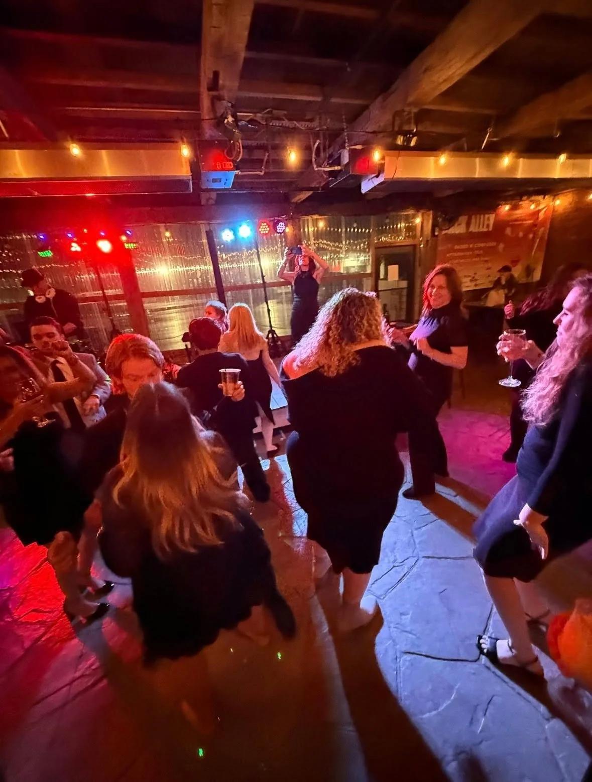 People dancing and socializing in a dimly lit bar or nightclub with colorful stage lights and a wooden ceiling.
