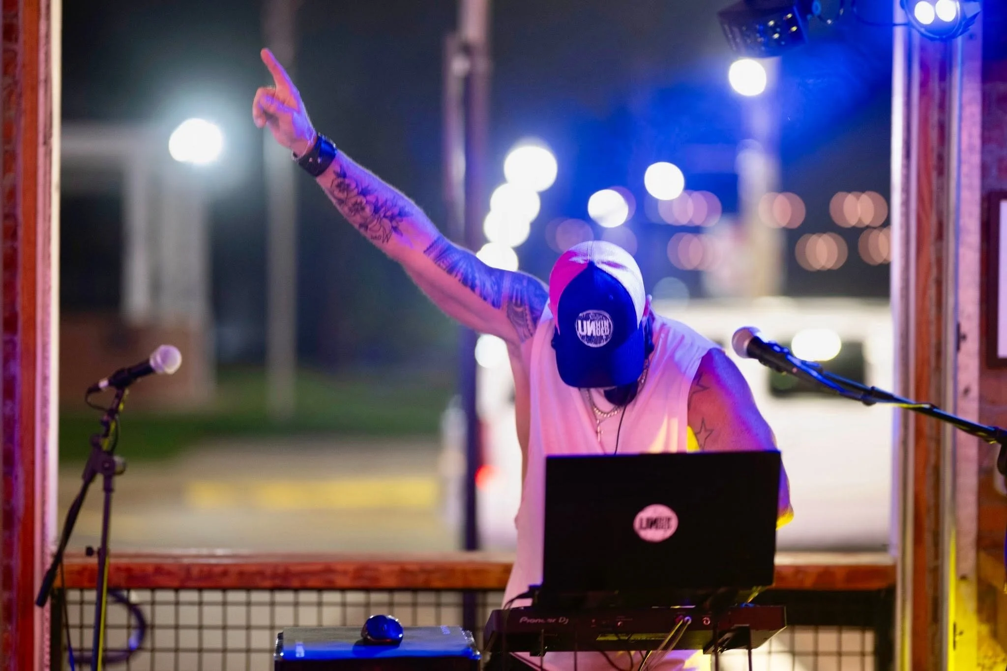 A person with tattoos on their arm, wearing a black and pink cap, DJing at an outdoor event at night. They are reaching out with their hand, and there are bright lights and a microphone in front of them.
