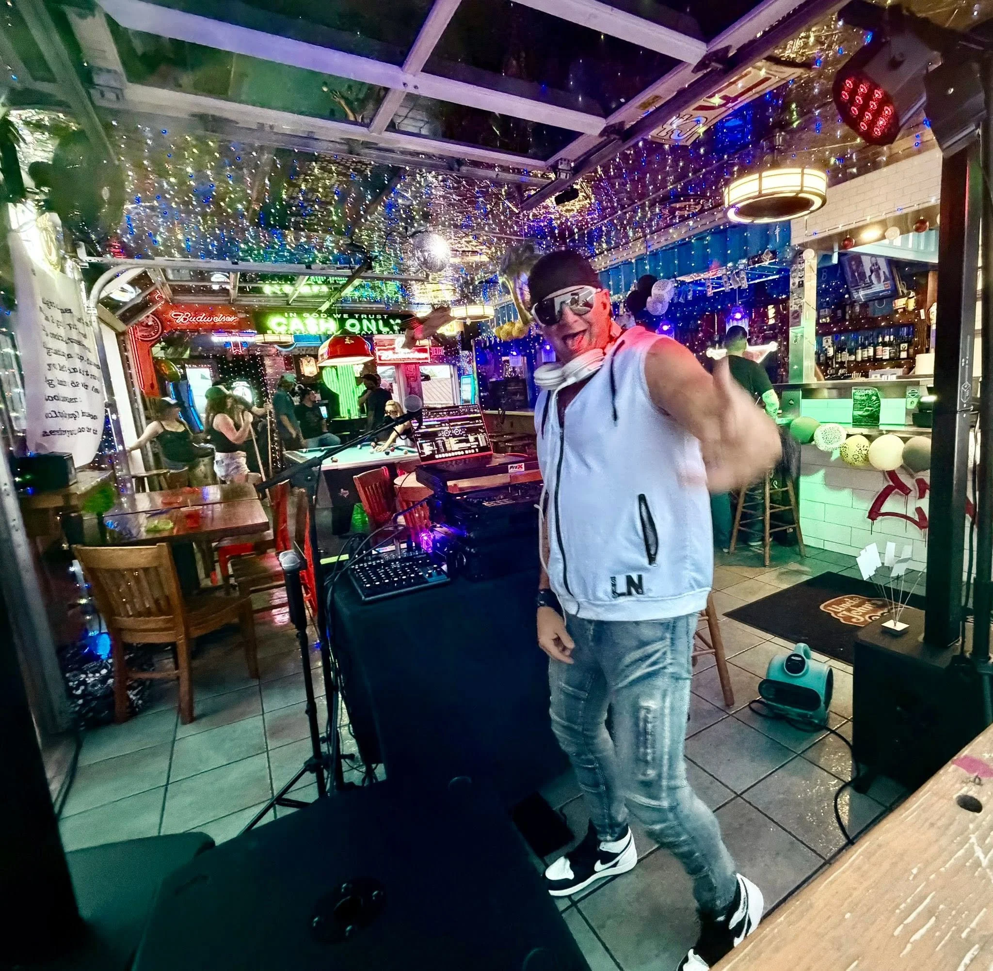 A person wearing glasses, a white sleeveless shirt, and jeans smiling and greeting a camera in a vibrant bar or nightclub decorated with colorful string lights. Others are seen in the background, some sitting at tables and others standing, under neon
