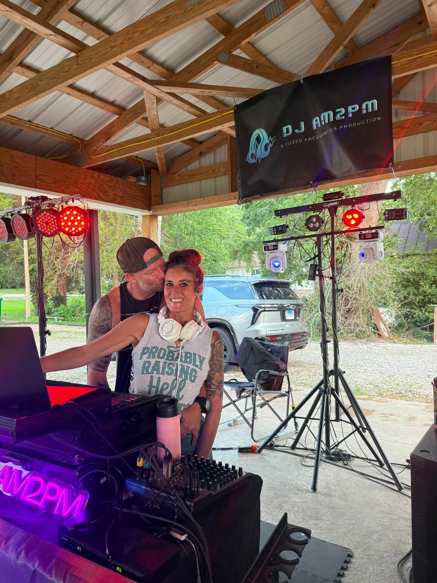 Two people, one man and one woman, in a wooden outdoor shelter with DJ and lighting equipment, trees and a vehicle in the background.