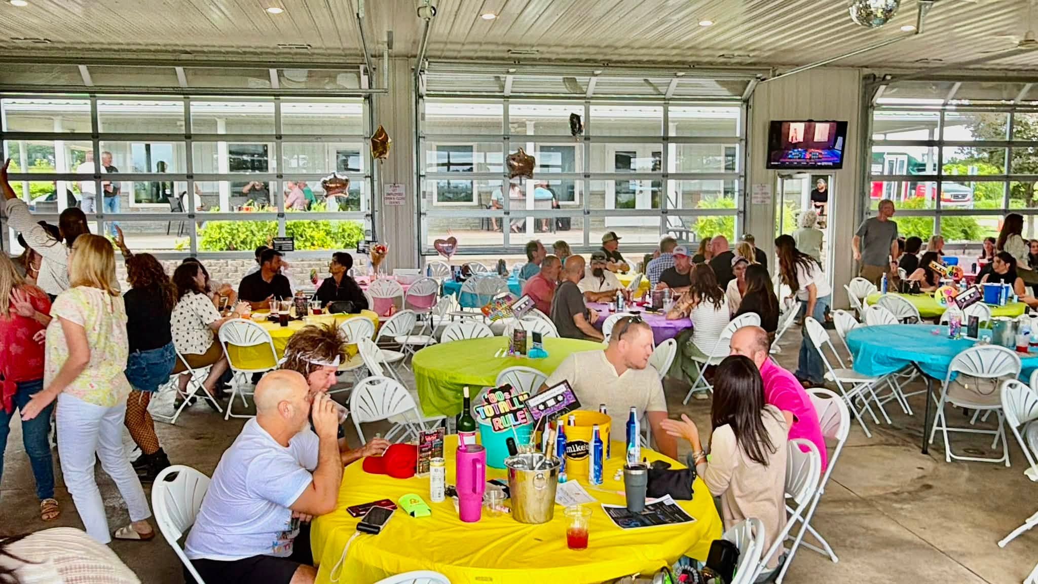 Indoor party with many people seated at decorated tables, balloons, and colorful tablecloths, large windows showing outside view, TV screen on the wall, some standing and mingling.