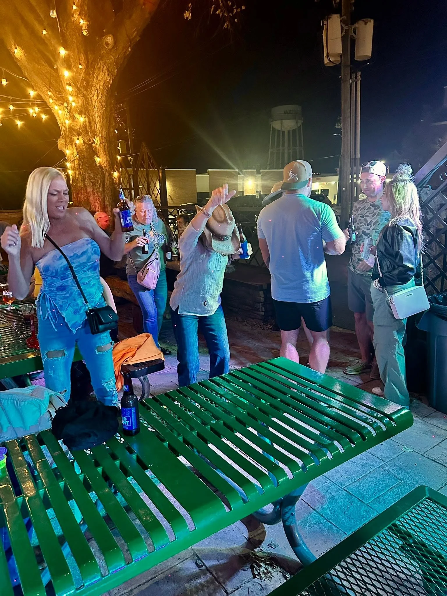 People socializing outdoors at night, holding drinks and talking, with a large tree decorated with string lights, a water tower visible in the background, and green benches in the foreground.